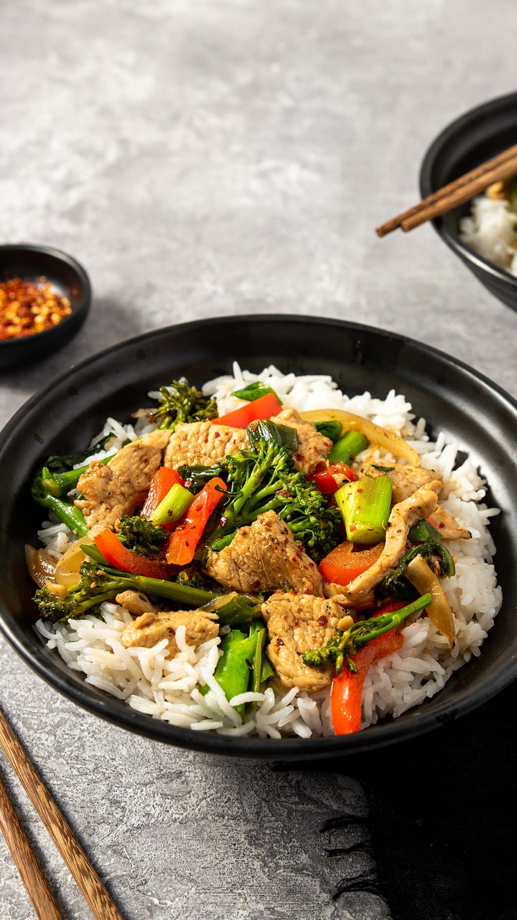 Asian stir-fry with colorful vegetables and crispy tofu served over white rice in a black bowl with chopsticks nearby.