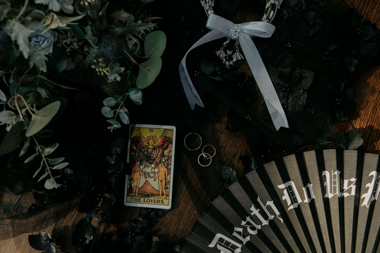 A tarot card and wedding rings arranged with greenery and a decorative fan on a dark wooden surface.
