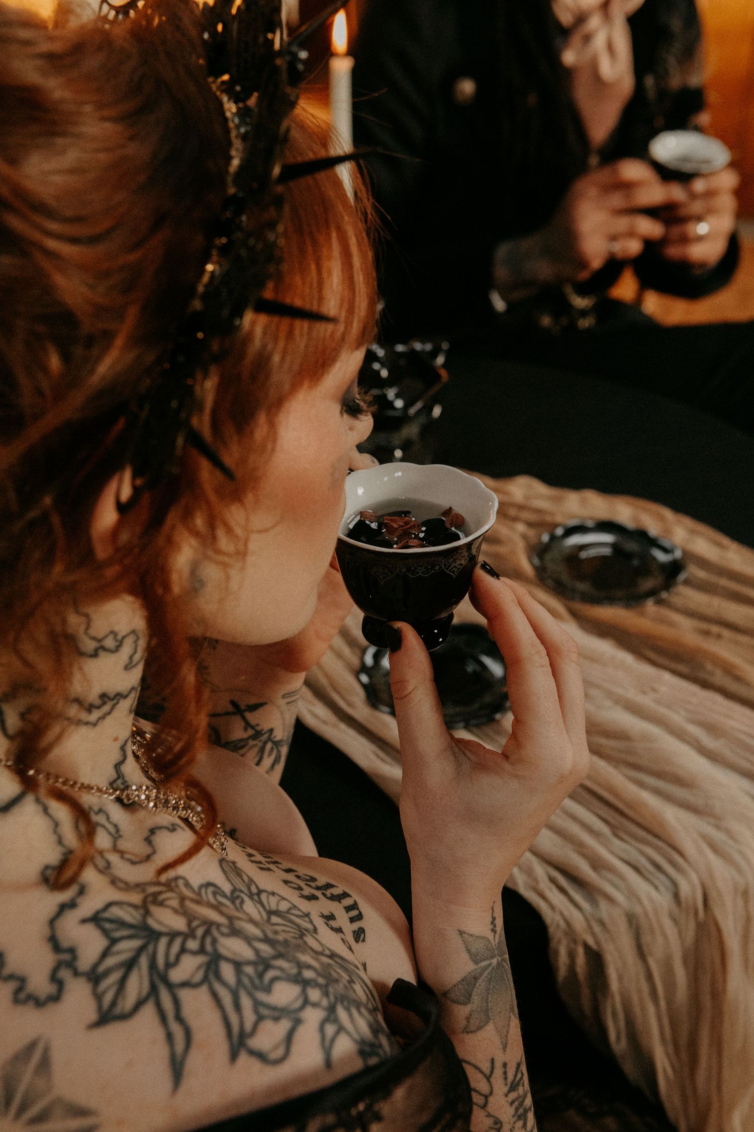 A heavily tattooed person holds a coffee cup while sitting at a wooden table in warm moody lighting.