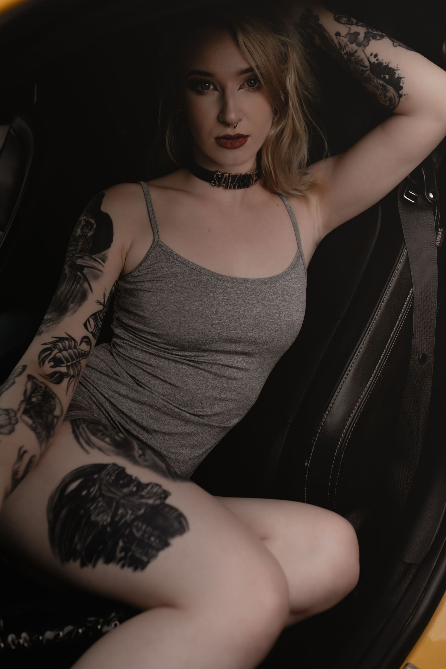 Moody boudoir photo of woman in gray tank top and tattoos sitting in car seat against dark background.