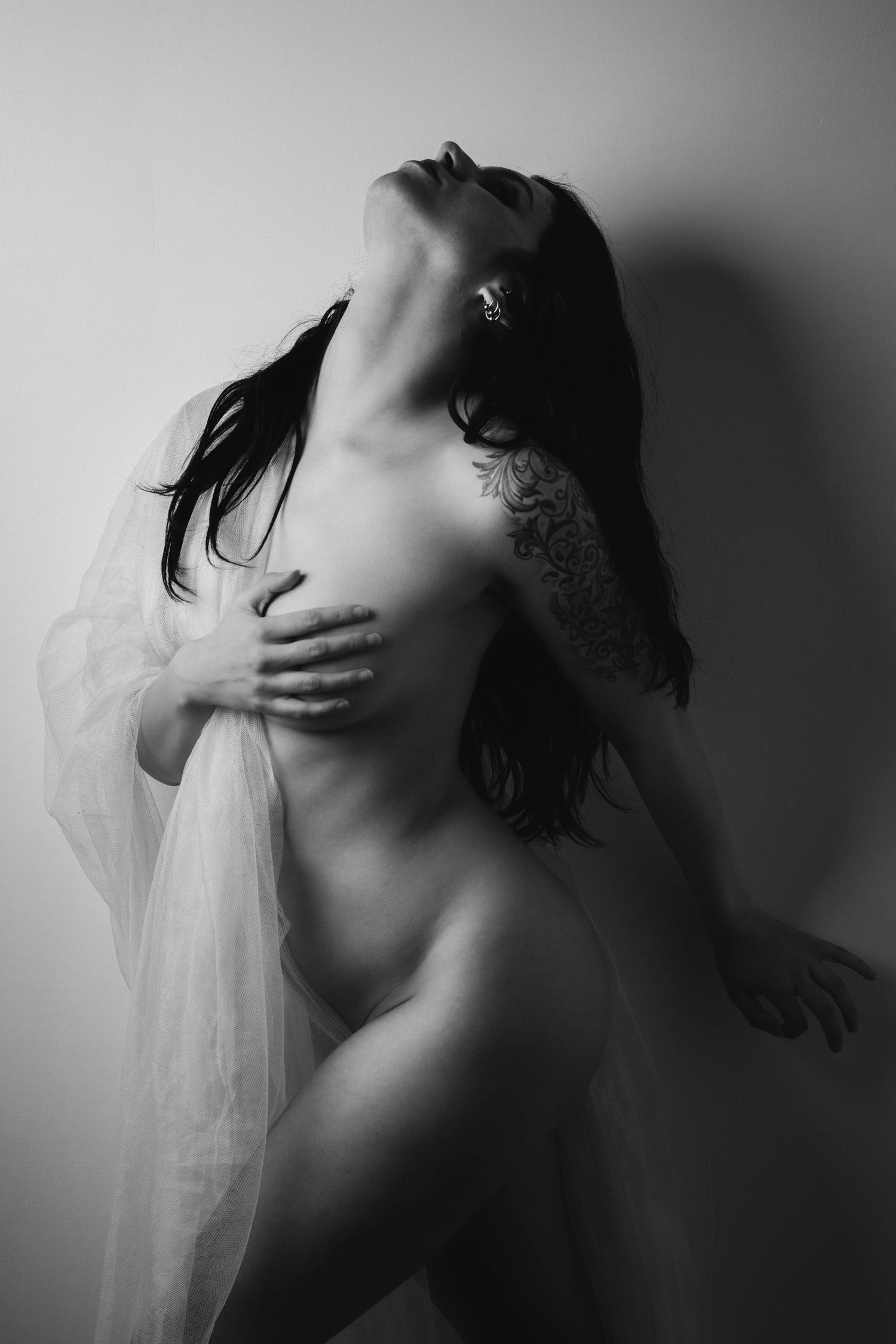 A monochromatic artistic nude silhouette in dramatic light holding sheer fabric with head tilted back.