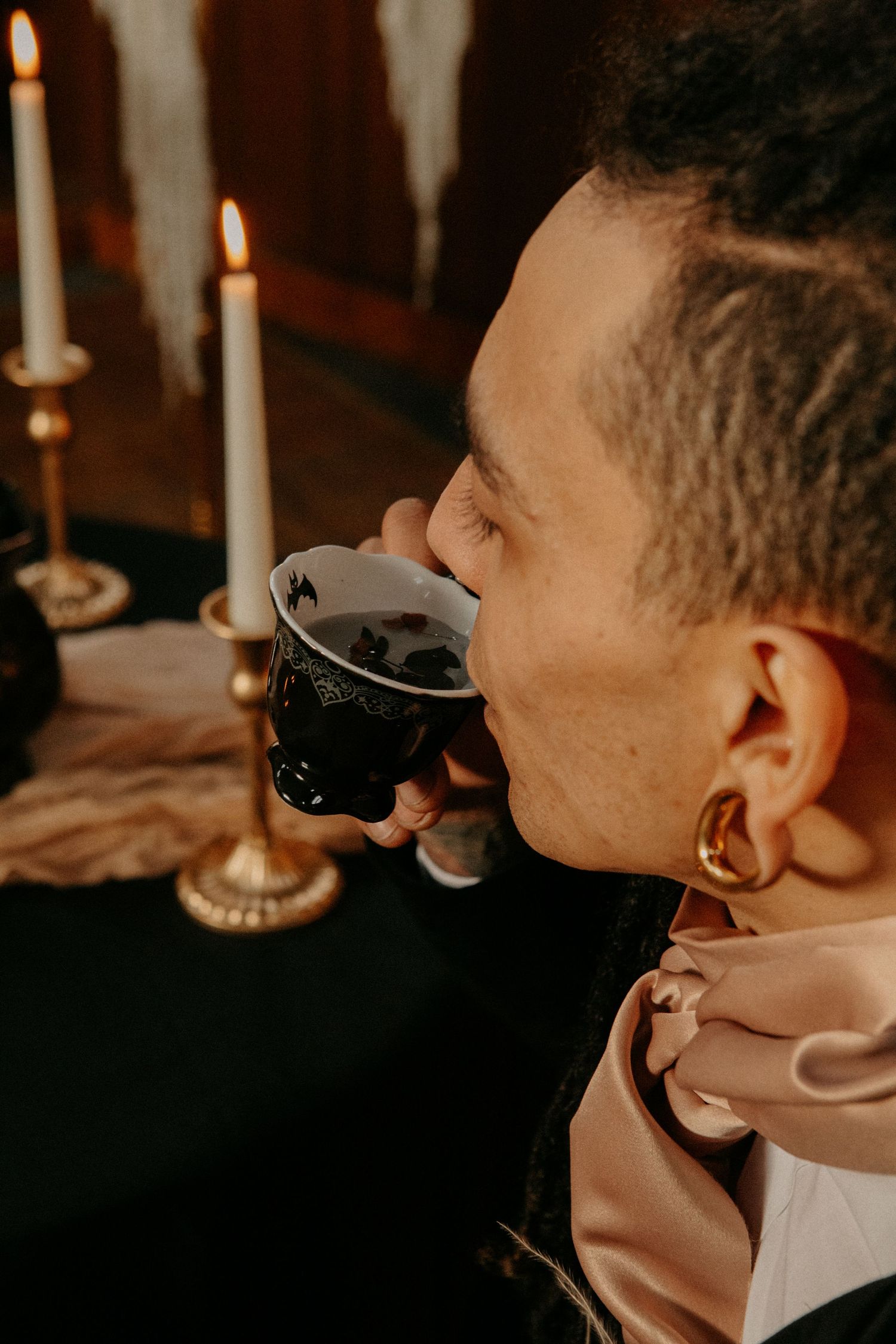 A person sipping from a dark mug by candlelight while wearing a beige top and earring in warm ambient lighting.
