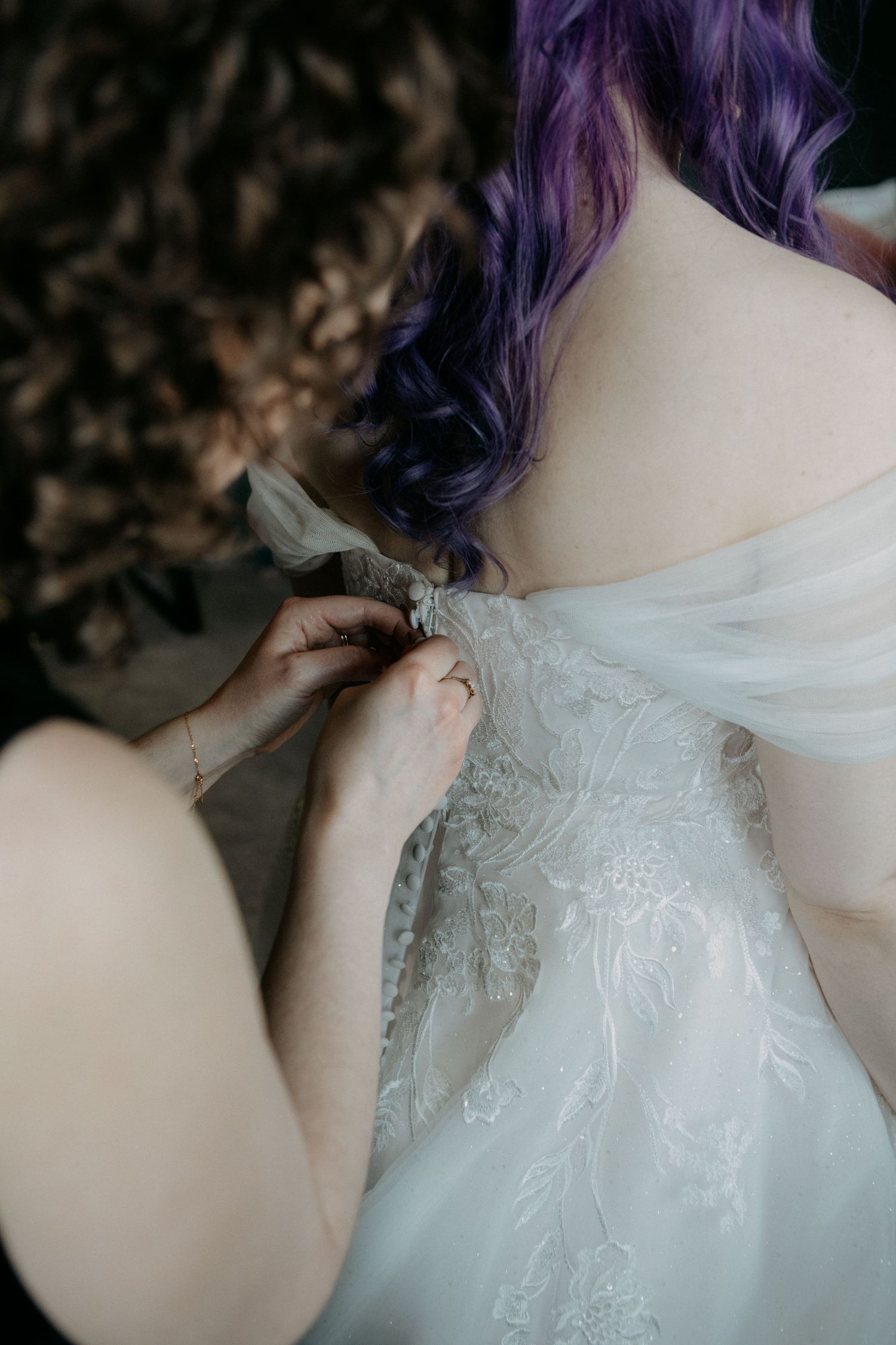 Close-up of delicate white lace wedding dress being fastened, with purple-dyed hair visible in soft focus.