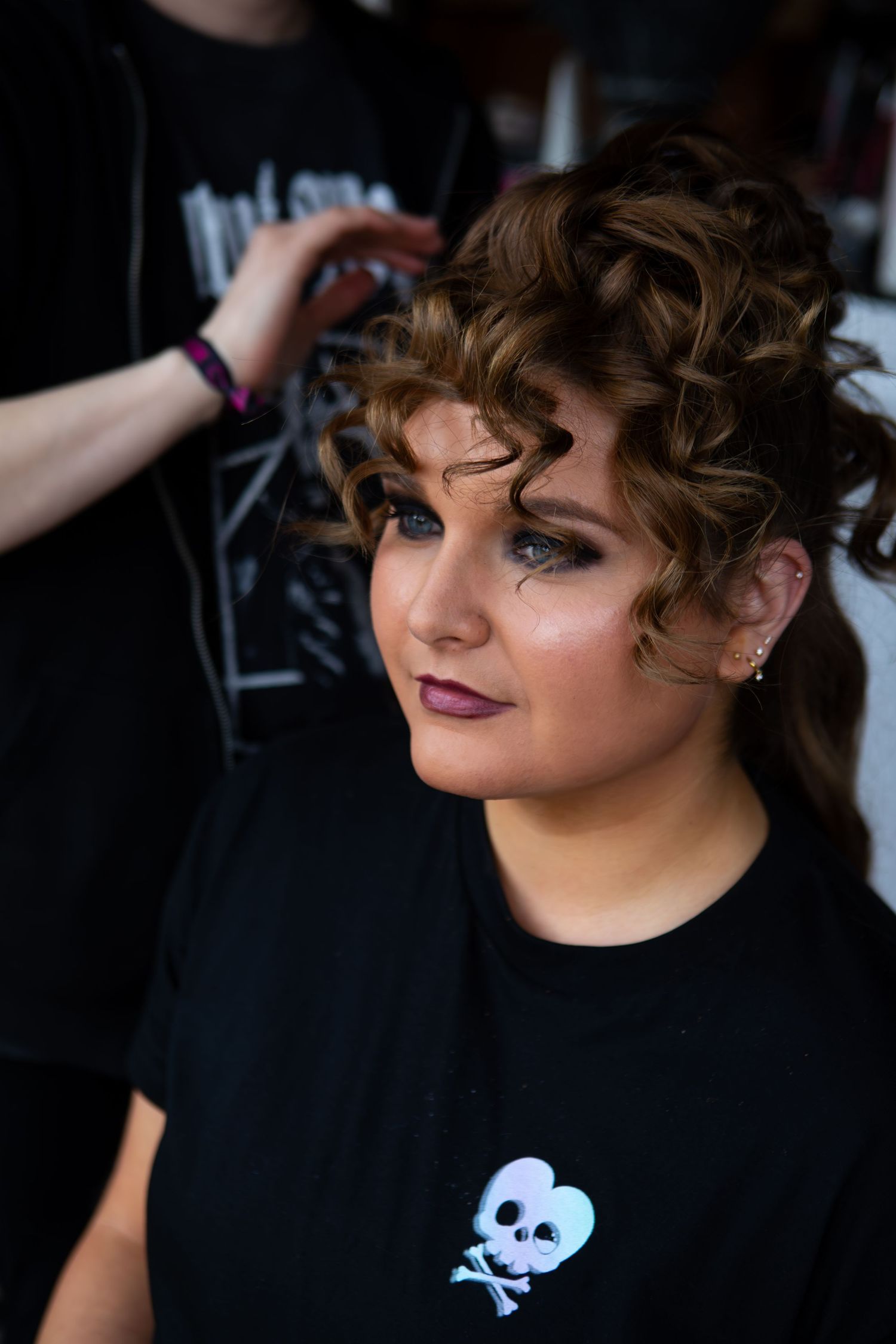 A person in a black t-shirt with skull graphics tilts their head while showing off dramatic eye makeup and curled hair.