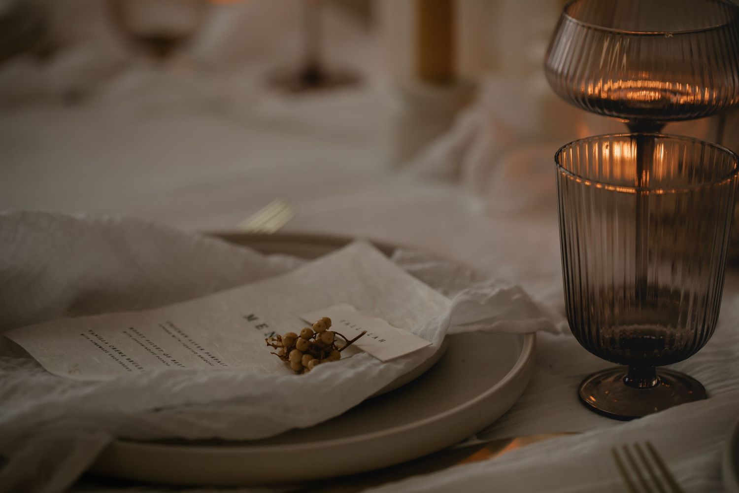 Moody table setting with a vintage glass and delicate dried flowers on white linen in warm candlelight.