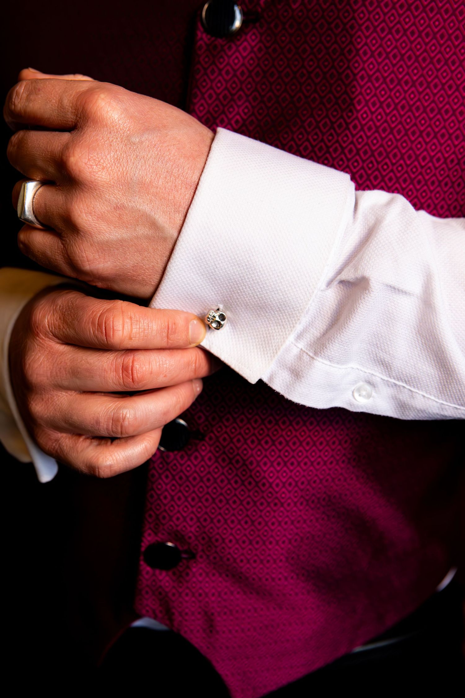 Close up of hands in a crisp white dress shirt adjusting a silver cufflink against a burgundy vest.