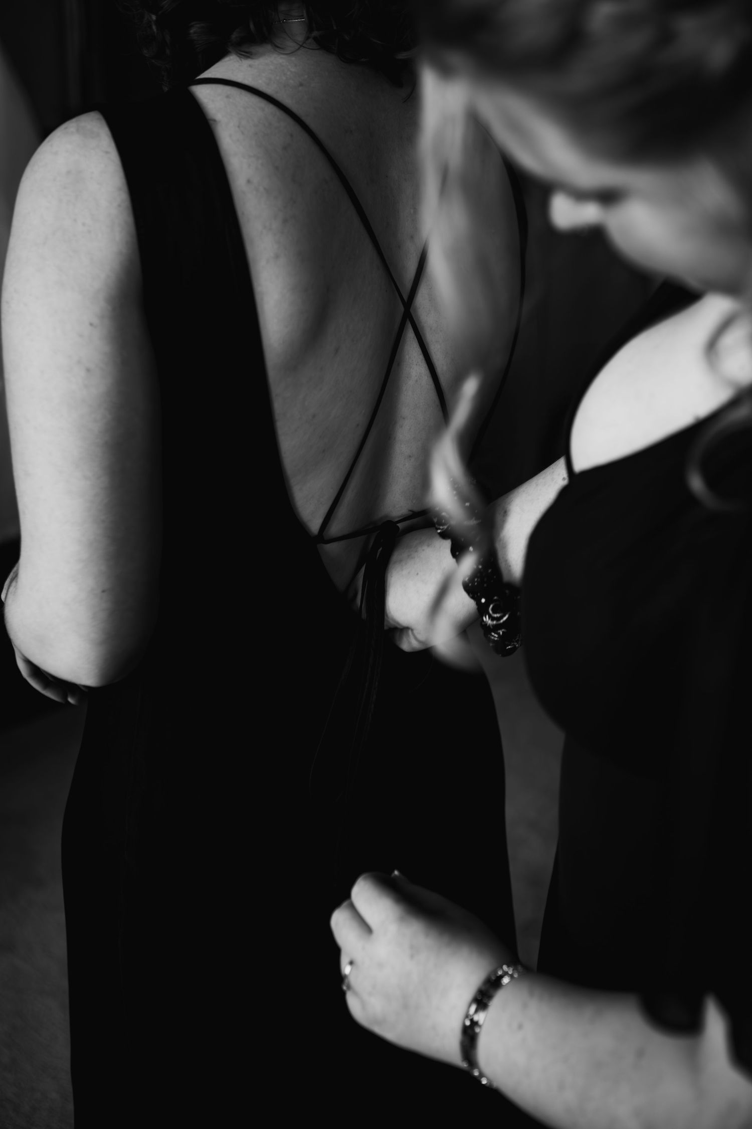 Black and white close-up of person in elegant backless dress being adjusted, showing straps and bracelet detail.