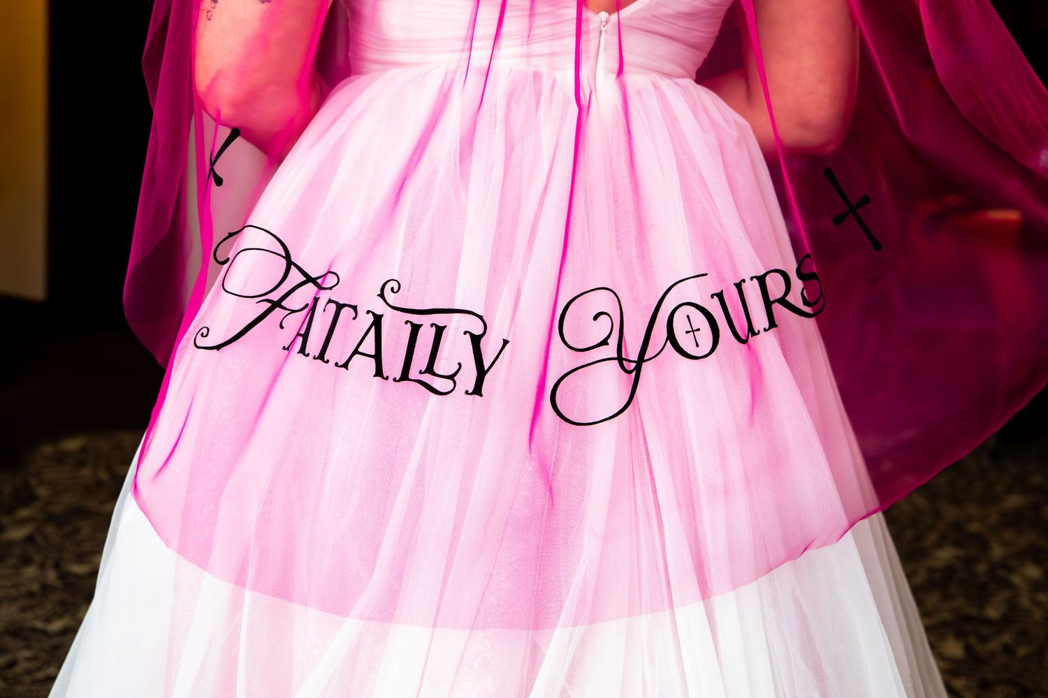 Pink bridal gown with elegant 'Fatally Yours' text embroidered on flowing fabric against dark background.