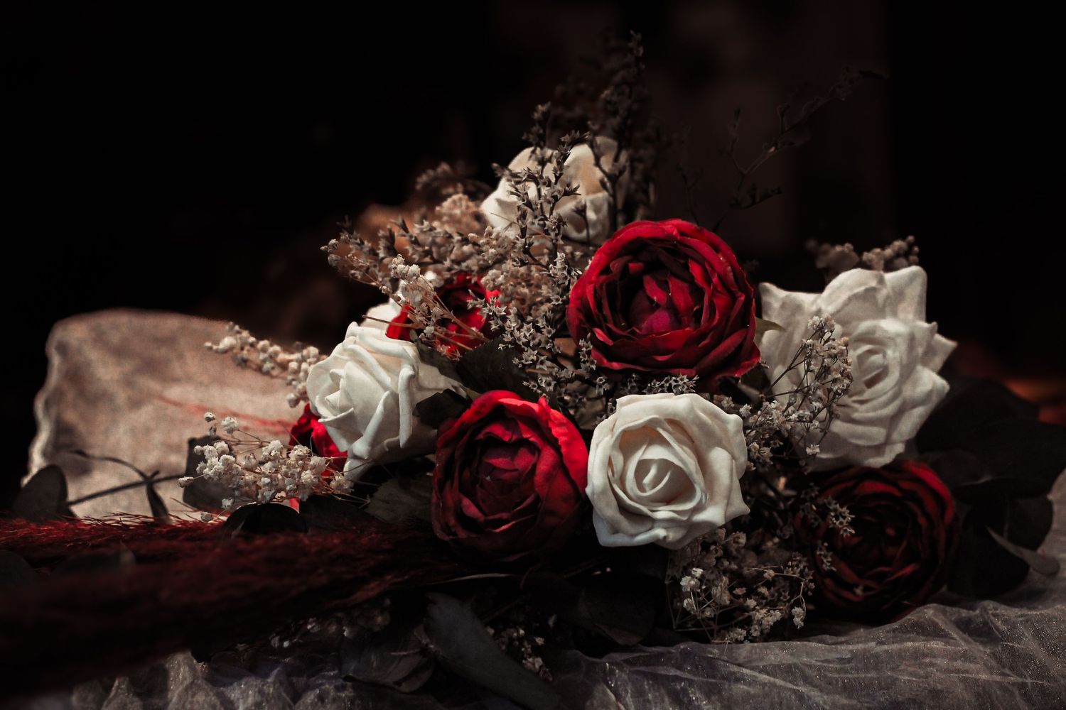Classic red and white roses bouquet in a dramatic dark setting with dried elements and fabric backdrop.