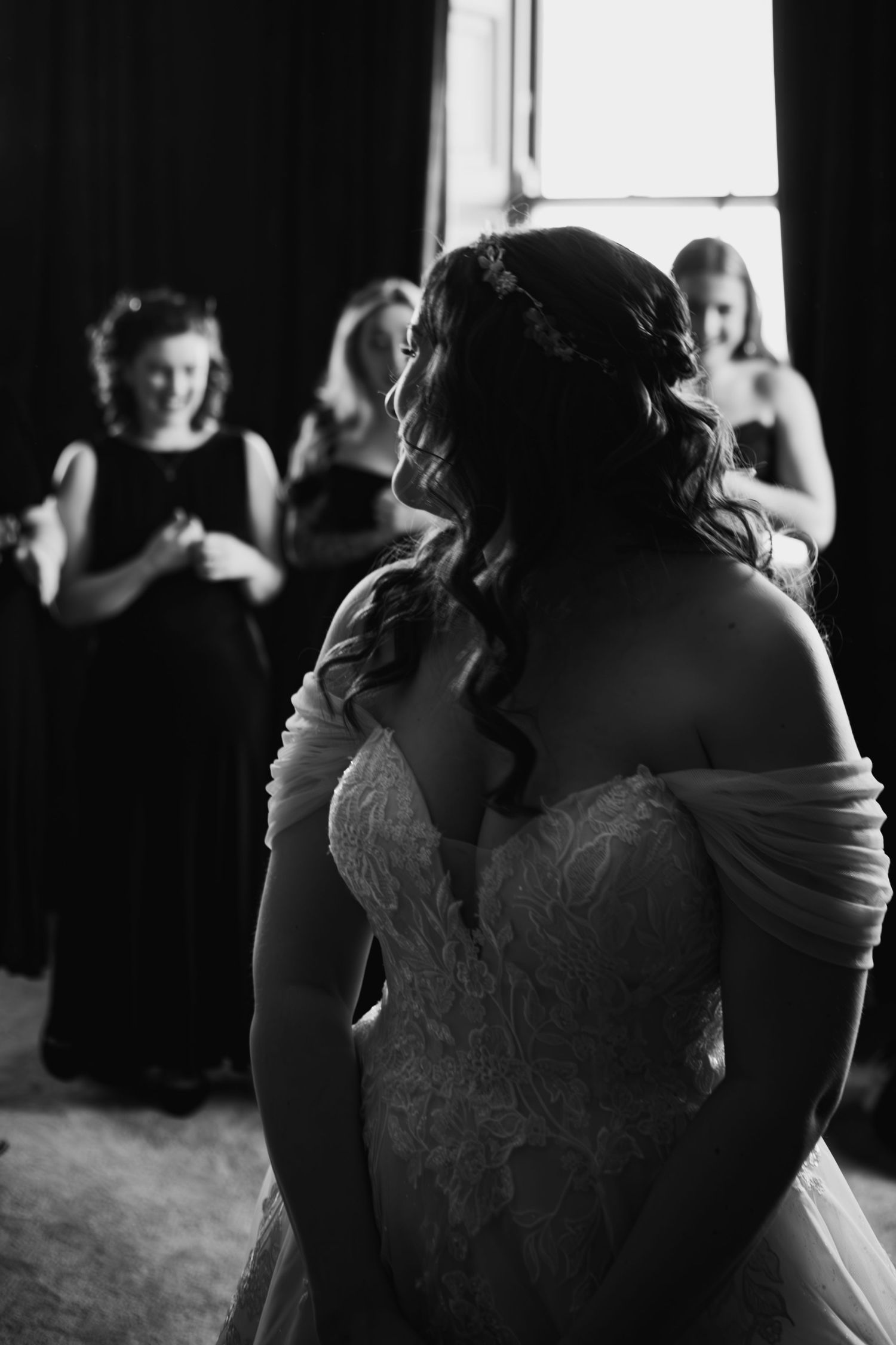 Black and white image showing a bride in a lace off-shoulder wedding gown with dark curled hair from behind.