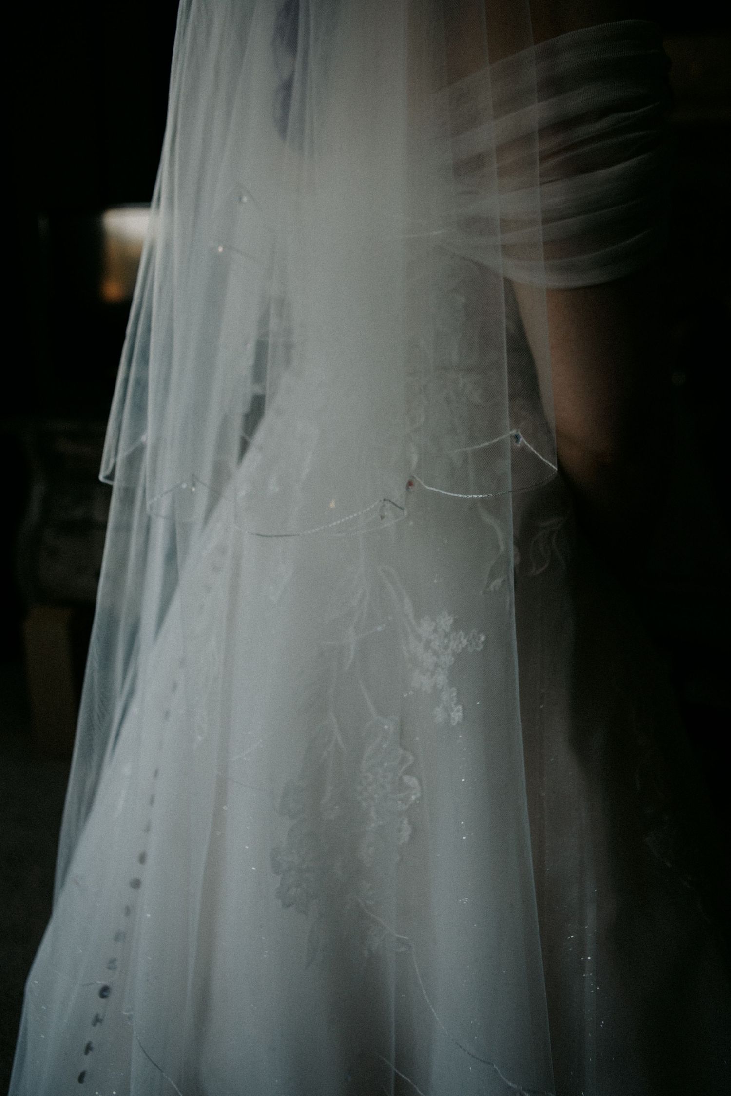 Close-up details of delicate white embroidery and lace patterns on a flowing wedding dress veil against a dark background.