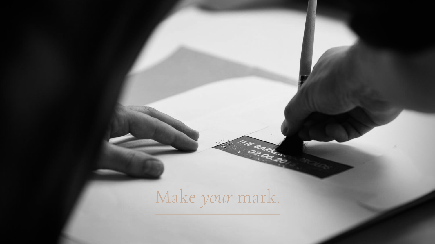 Close-up black and white view of hands writing on paper with a pen showing 'make your mark' text.