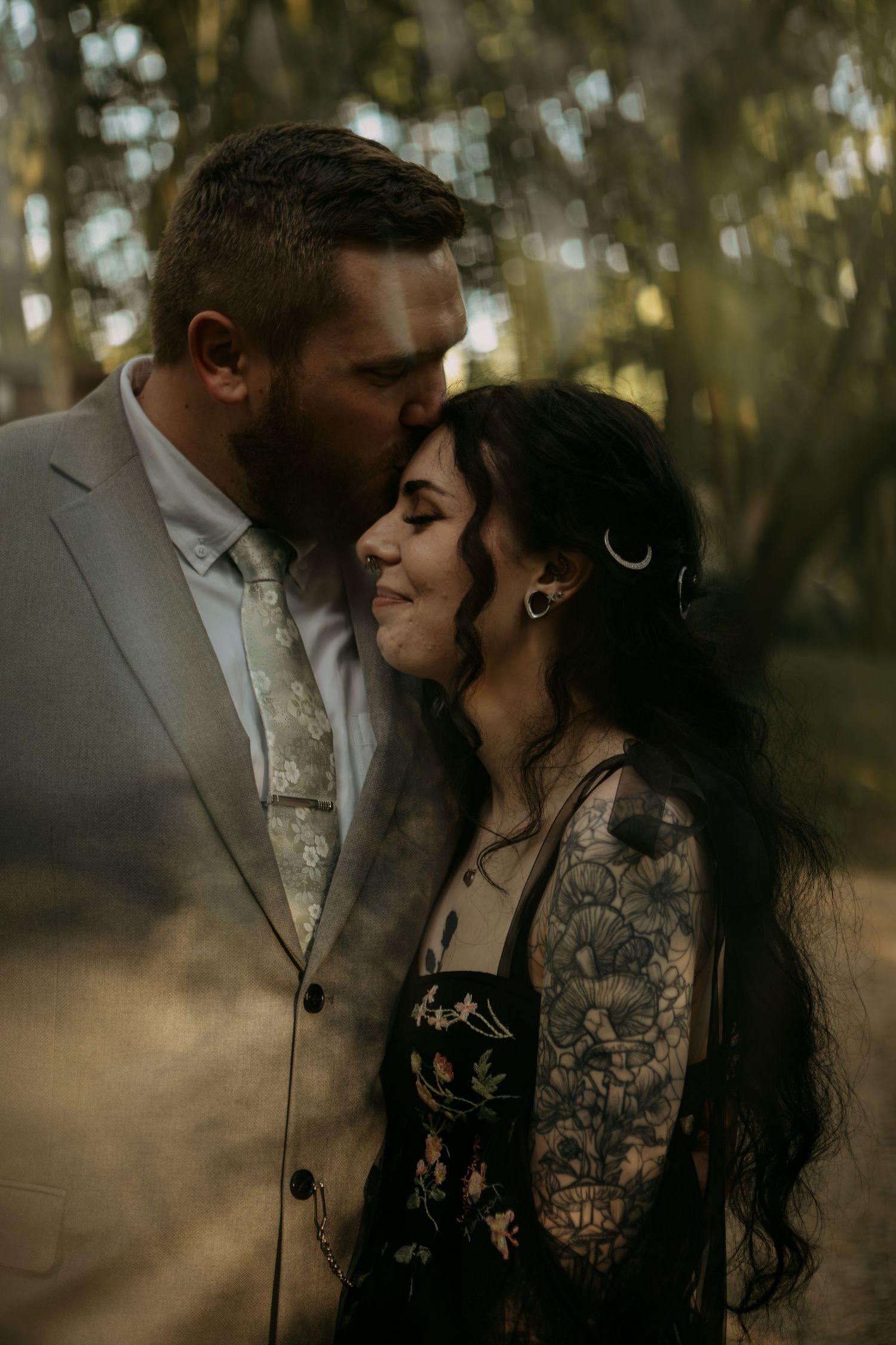 A couple shares an intimate moment in a forest setting, with soft light filtering through the trees.
