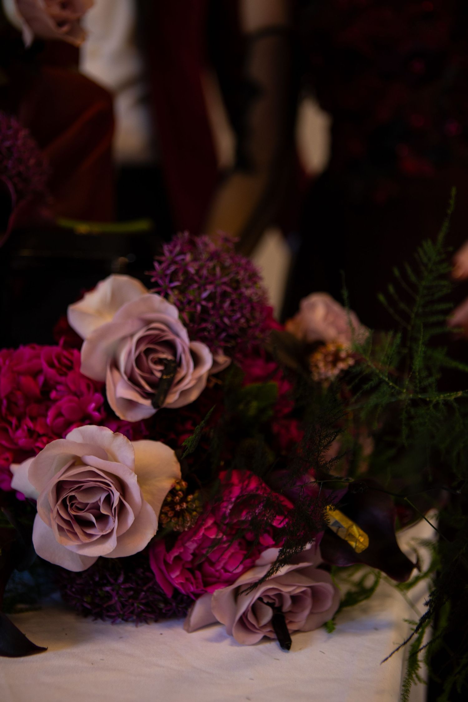 Moody floral arrangement featuring soft pink roses and deep burgundy flowers with delicate ferns on white surface.