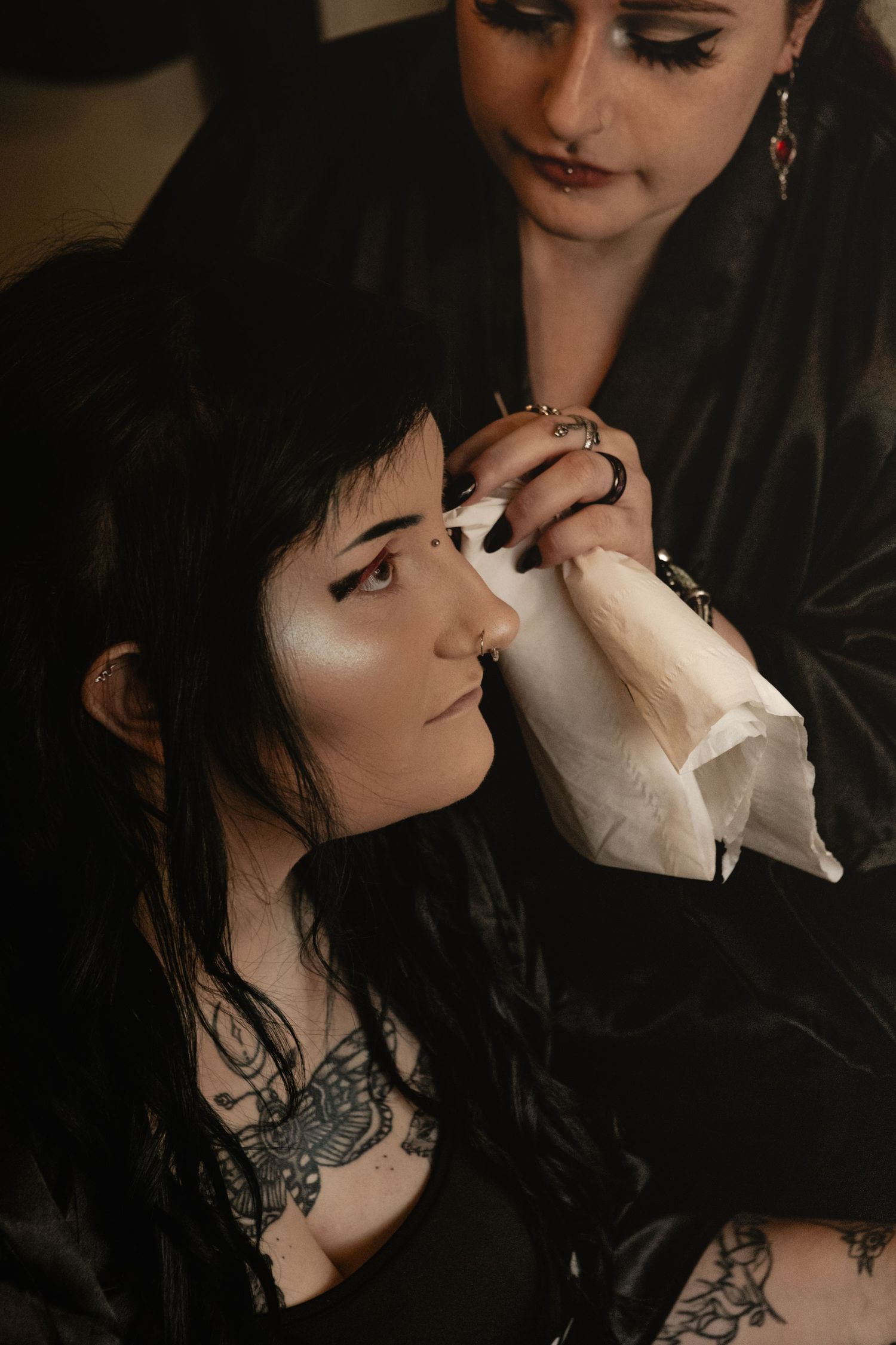 Makeup artist applying dramatic eye makeup in moody lighting with gothic aesthetic details and visible tattoos.