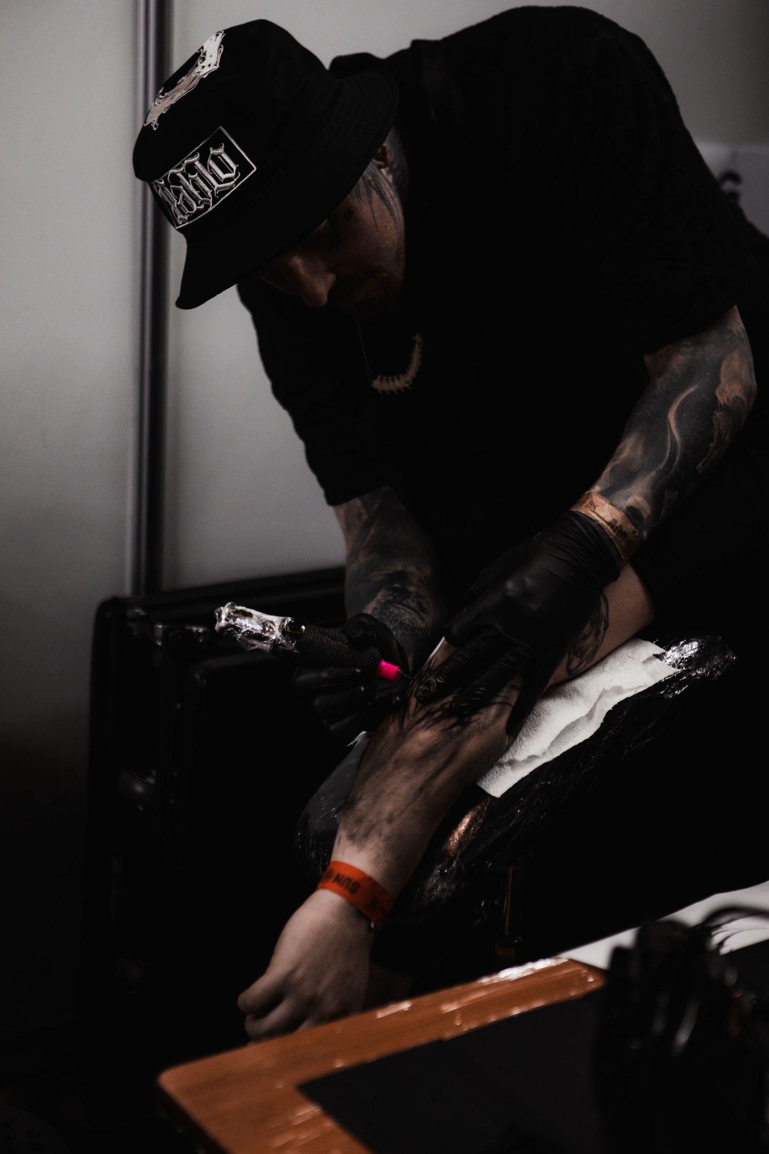 A tattoo artist wearing dark clothing works focused on a client in a dimly lit studio setting.