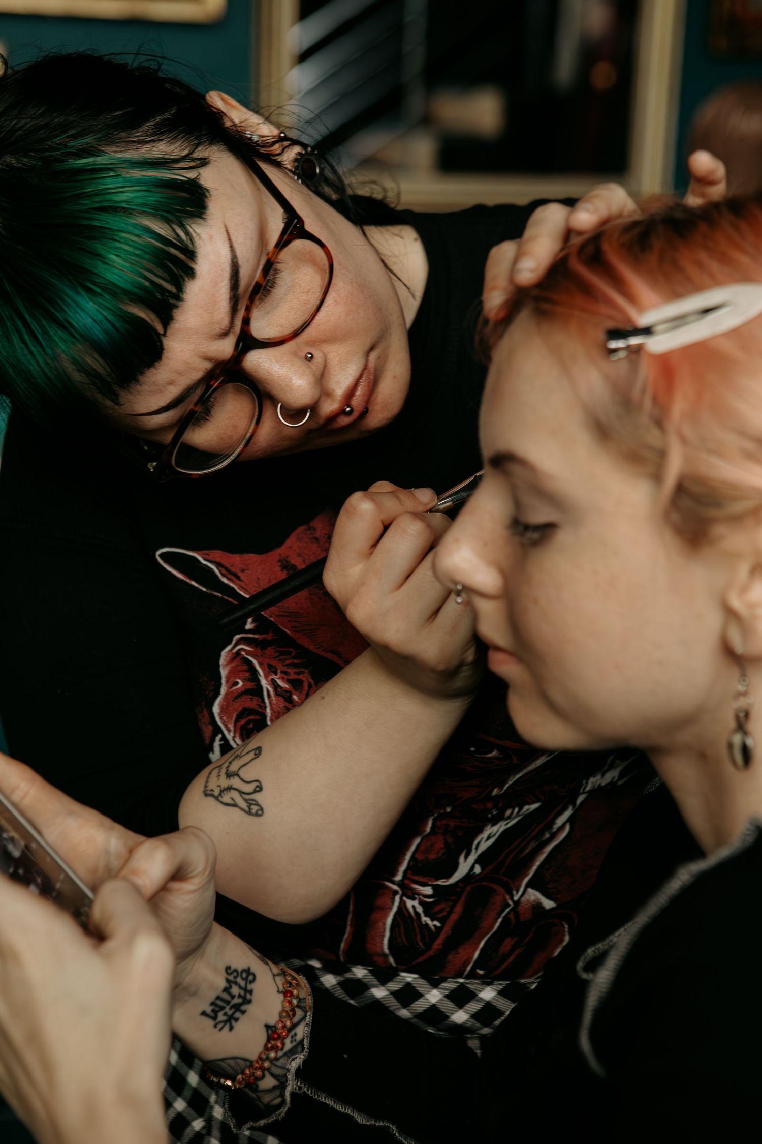 Two people with colorful dyed hair share a moment as one applies makeup to the other in a close-up intimate setting.
