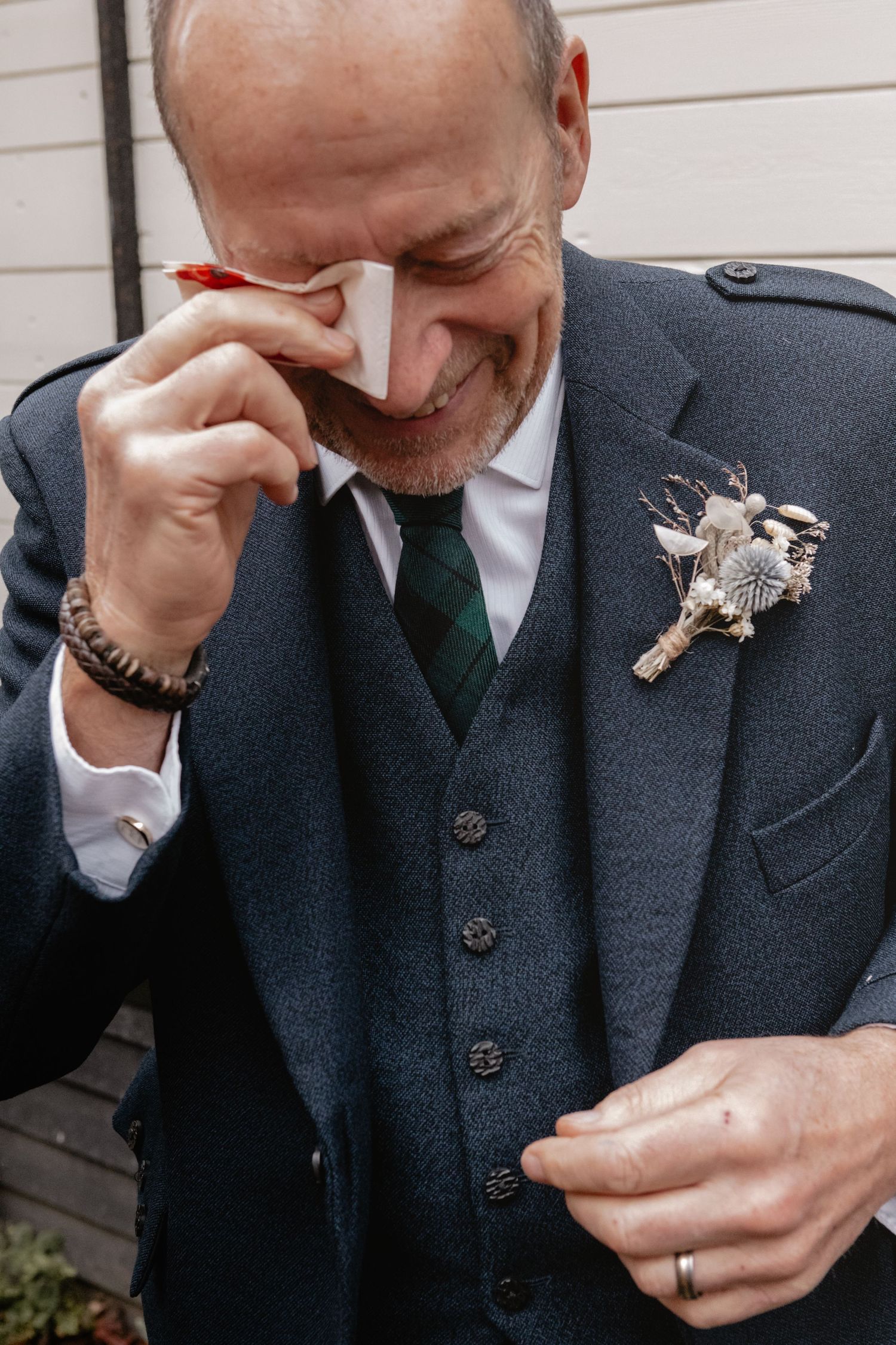 Emotional groom in navy three-piece suit wipes tears with handkerchief while wearing a unique floral boutonniere.