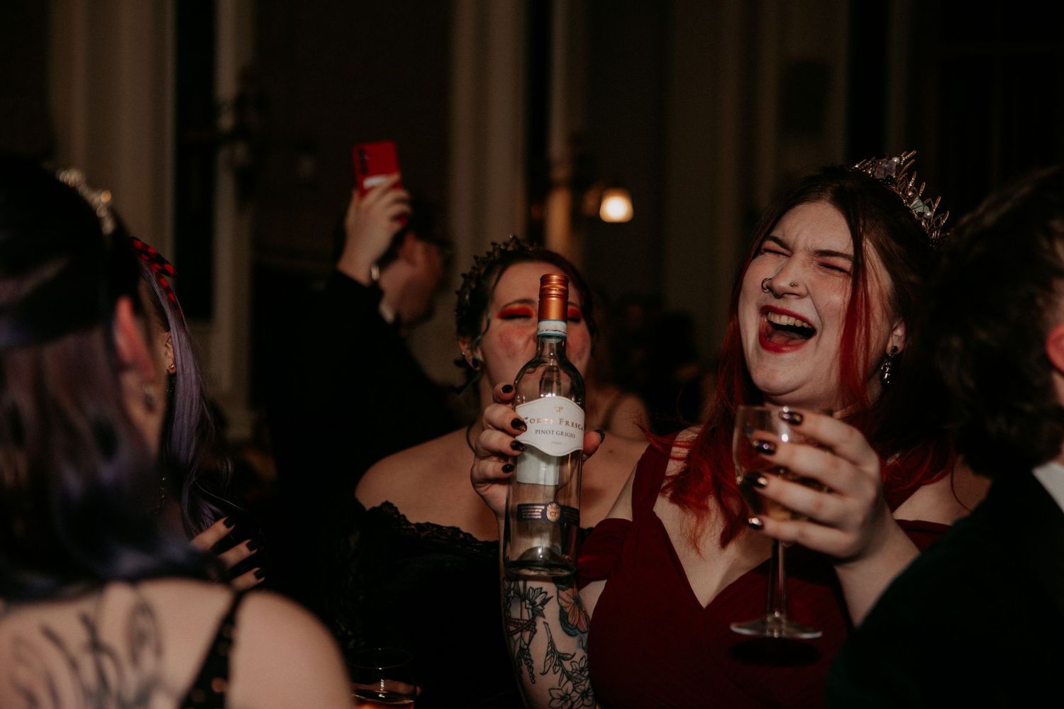 Party guests in dark attire enjoying drinks and laughing together in a dimly lit setting.