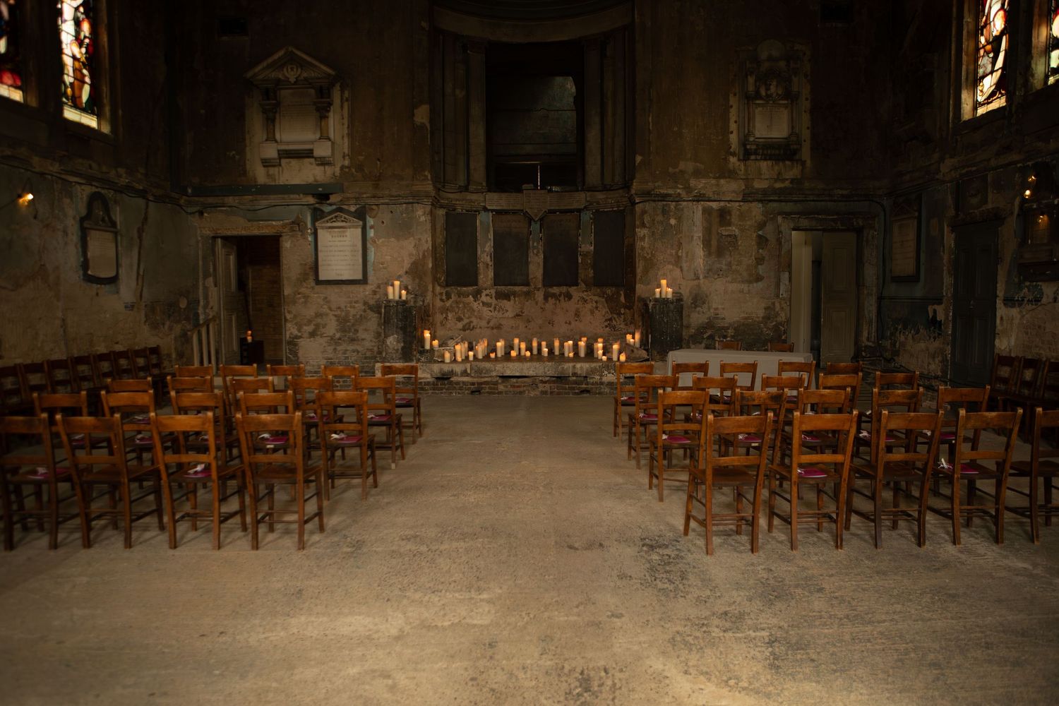 Empty rows of wooden chairs face an altar in a dimly lit, historic stone chapel with stained glass windows.