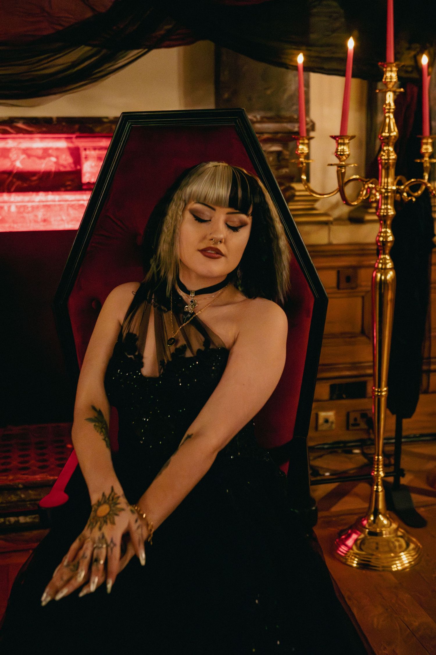 A gothic scene with a velvet-lined coffin, golden candelabra, and dark dress against dramatic red lighting.