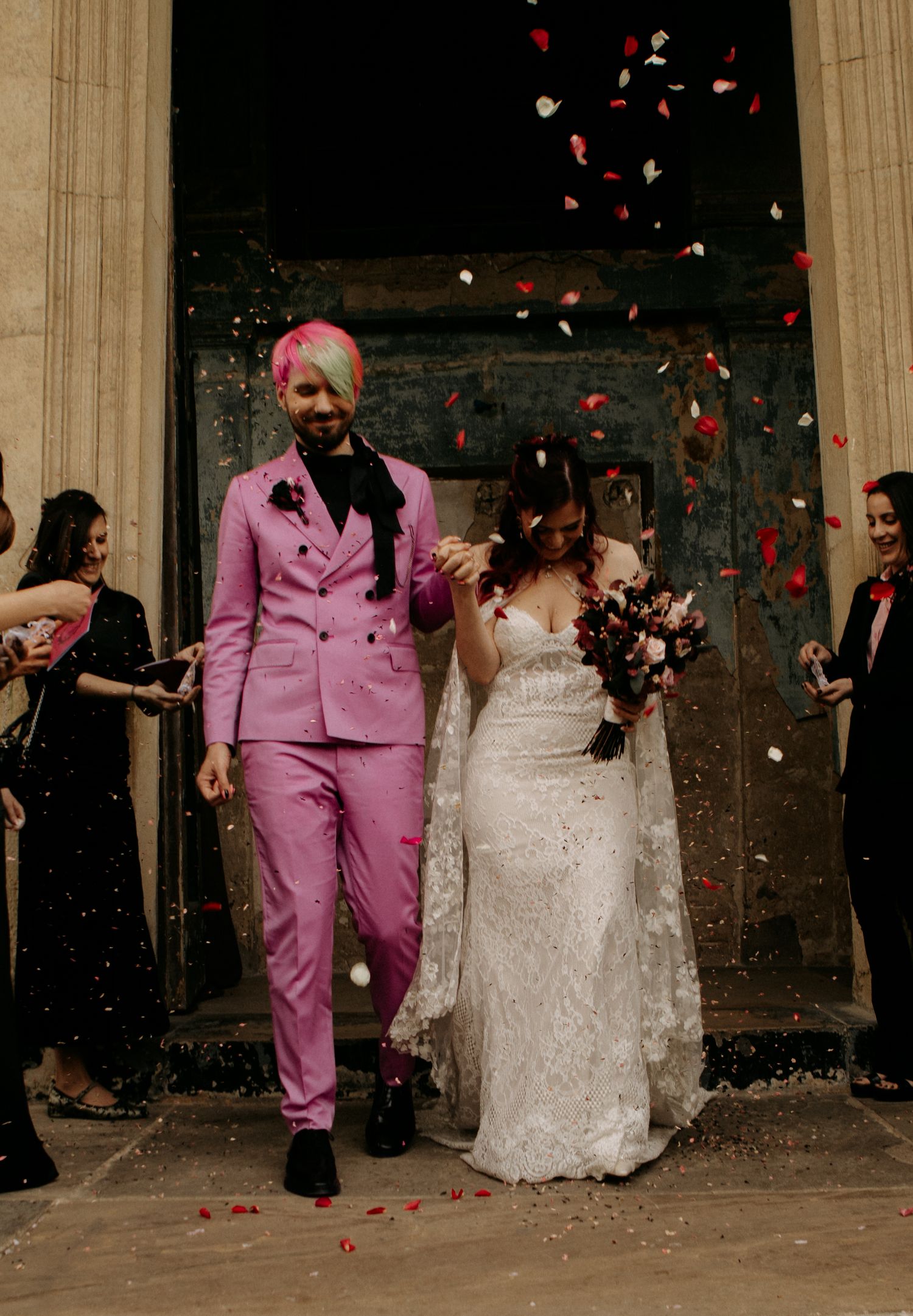 Wedding couple exits church under shower of red rose petals with one wearing striking pink suit and another in white lace gown.