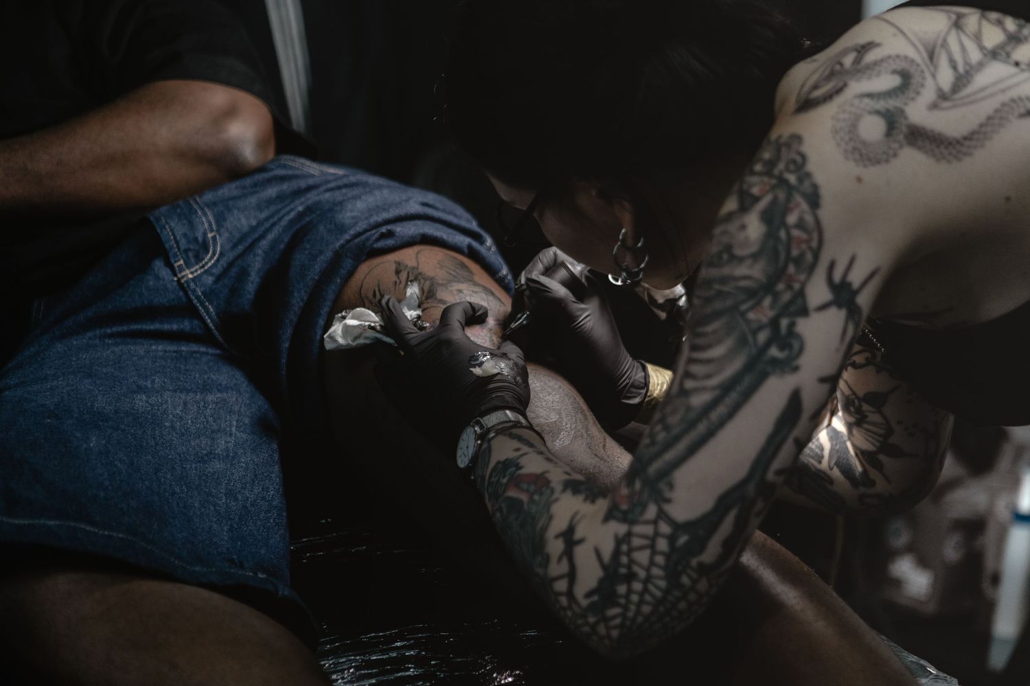 A tattoo artist works on a detailed design while wearing protective gloves in a dimly lit studio.