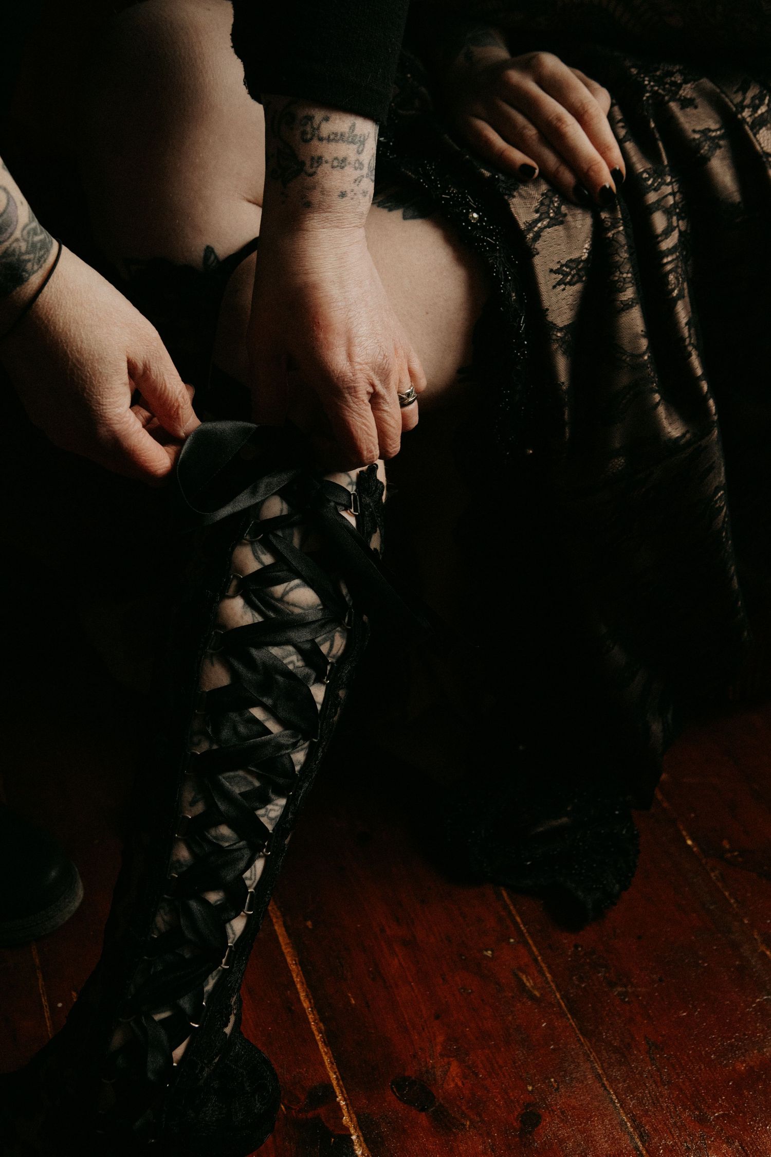 Hands lacing up a black corset-style boot on a wooden floor in moody lighting.
