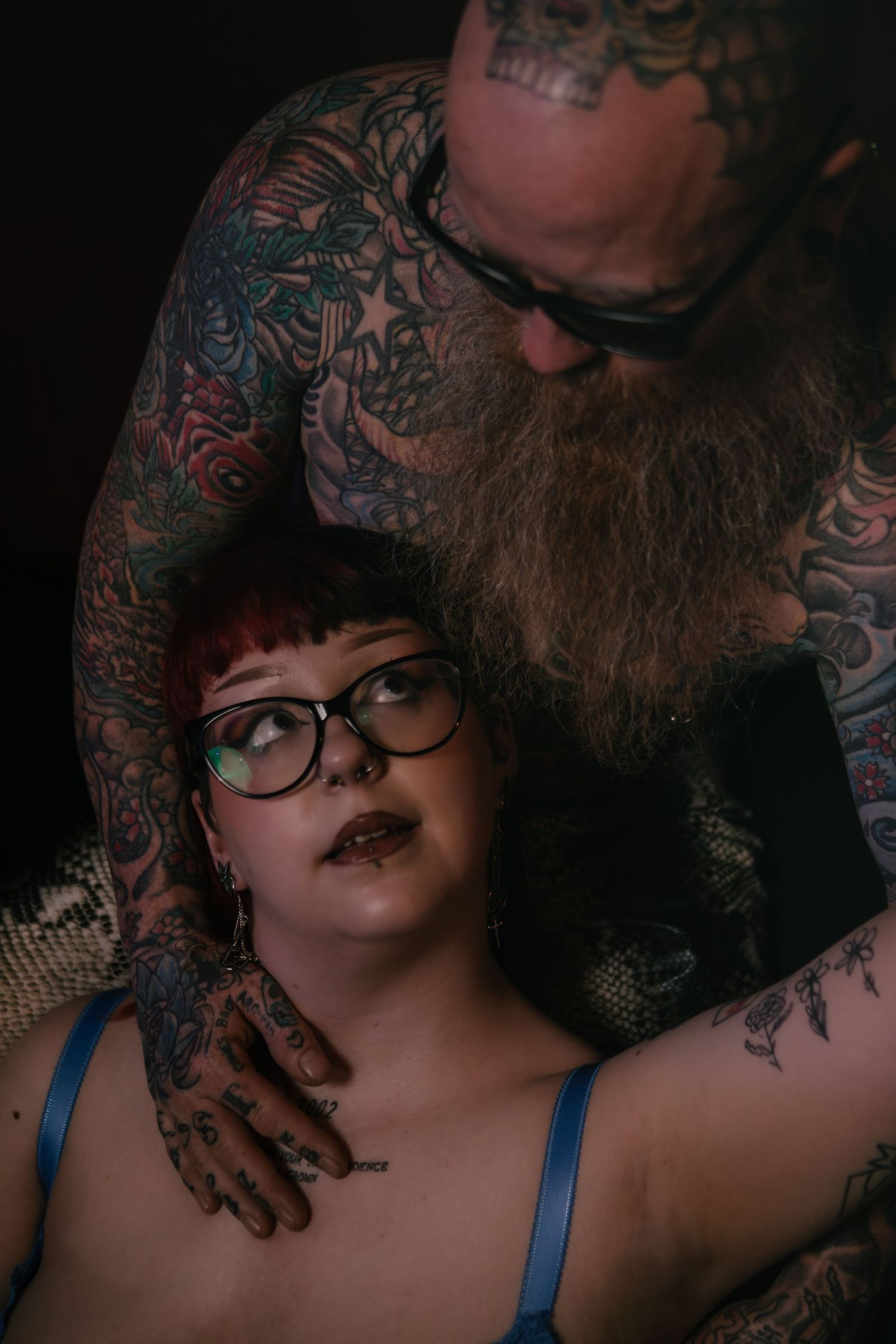 A moody boudoir portrait featuring body art, showing an intimate couple with extensive tattoos and dark ambiance.
