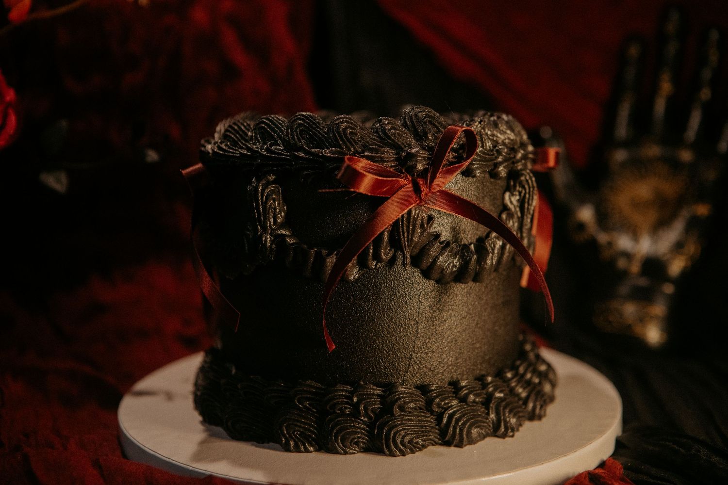 A dark chocolate cake with black frosted rosettes and red ribbon sits on a white plate against a dramatic red background.