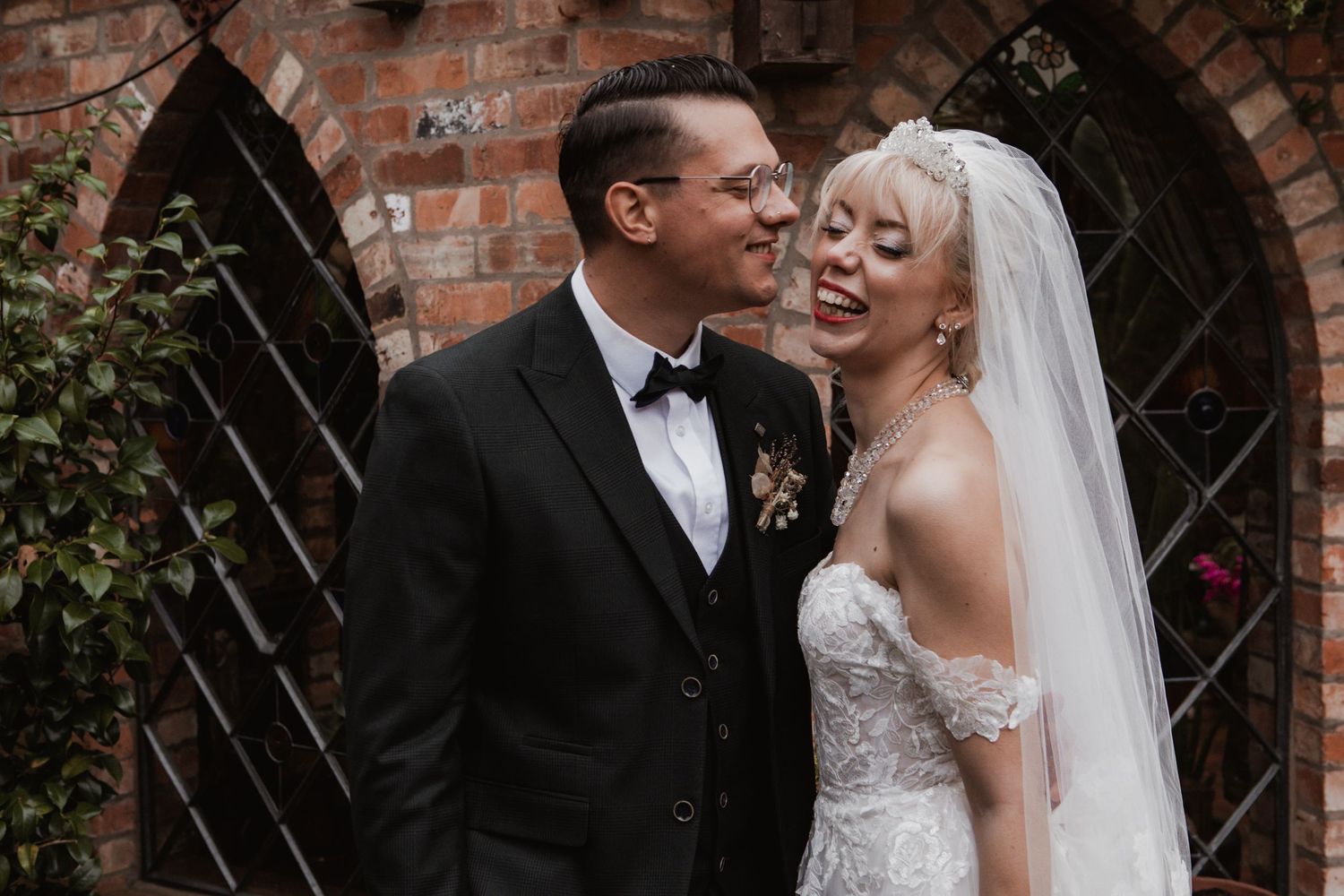 A romantic wedding couple poses against a rustic brick wall background.