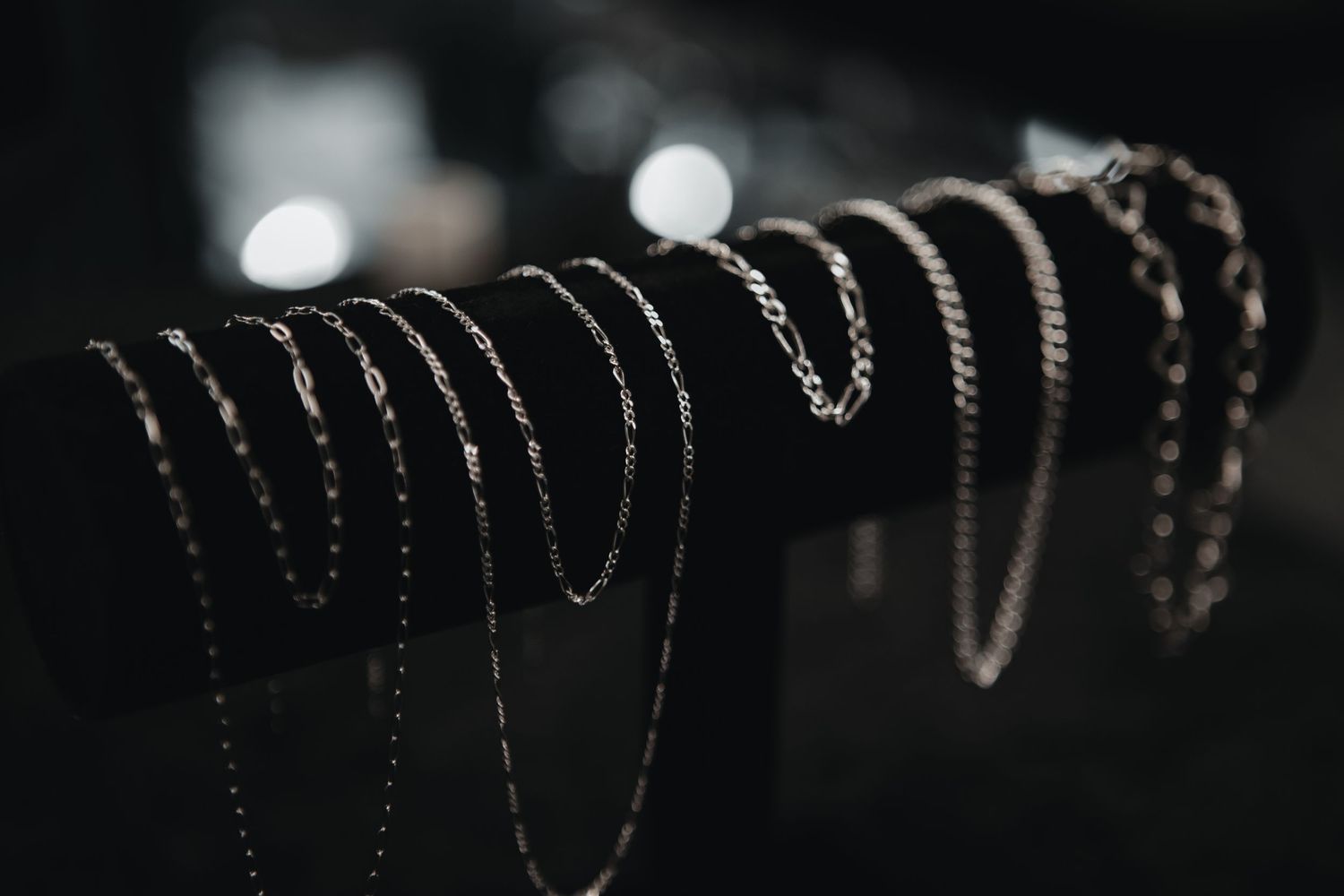 Multiple delicate silver necklace chains draped over a black display bar with soft bokeh lights in the background.