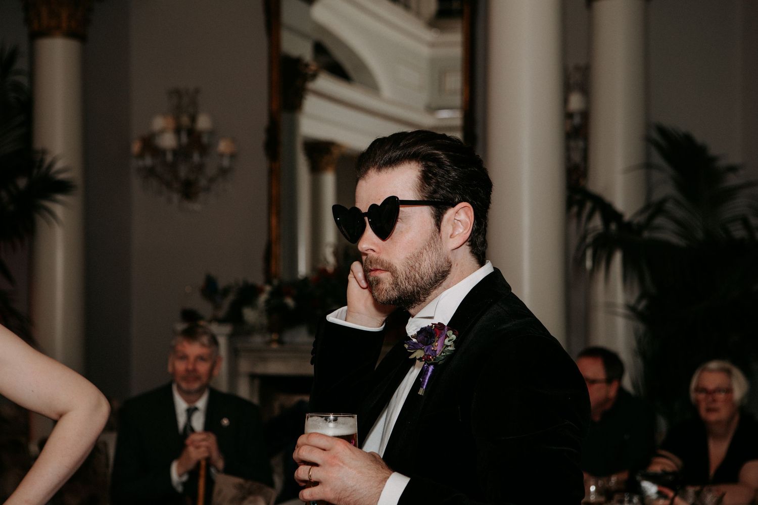 Wedding guest in dark suit and sunglasses sips a drink while standing in an elegant venue with columns.