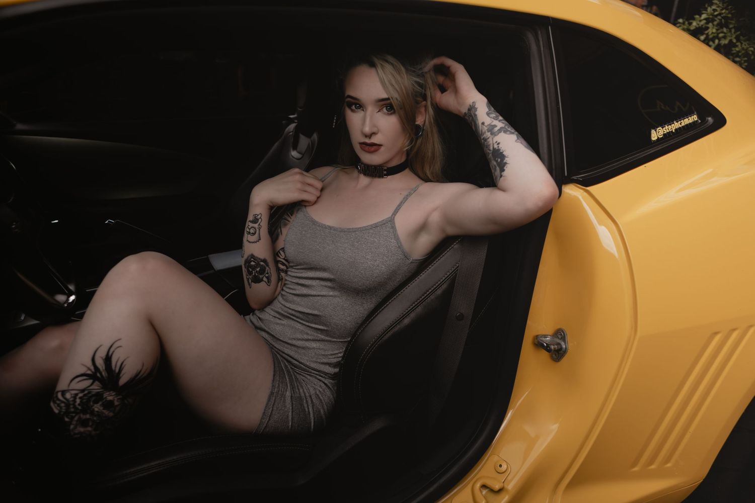 A person poses elegantly in a grey dress inside a bright yellow luxury sports car at night.