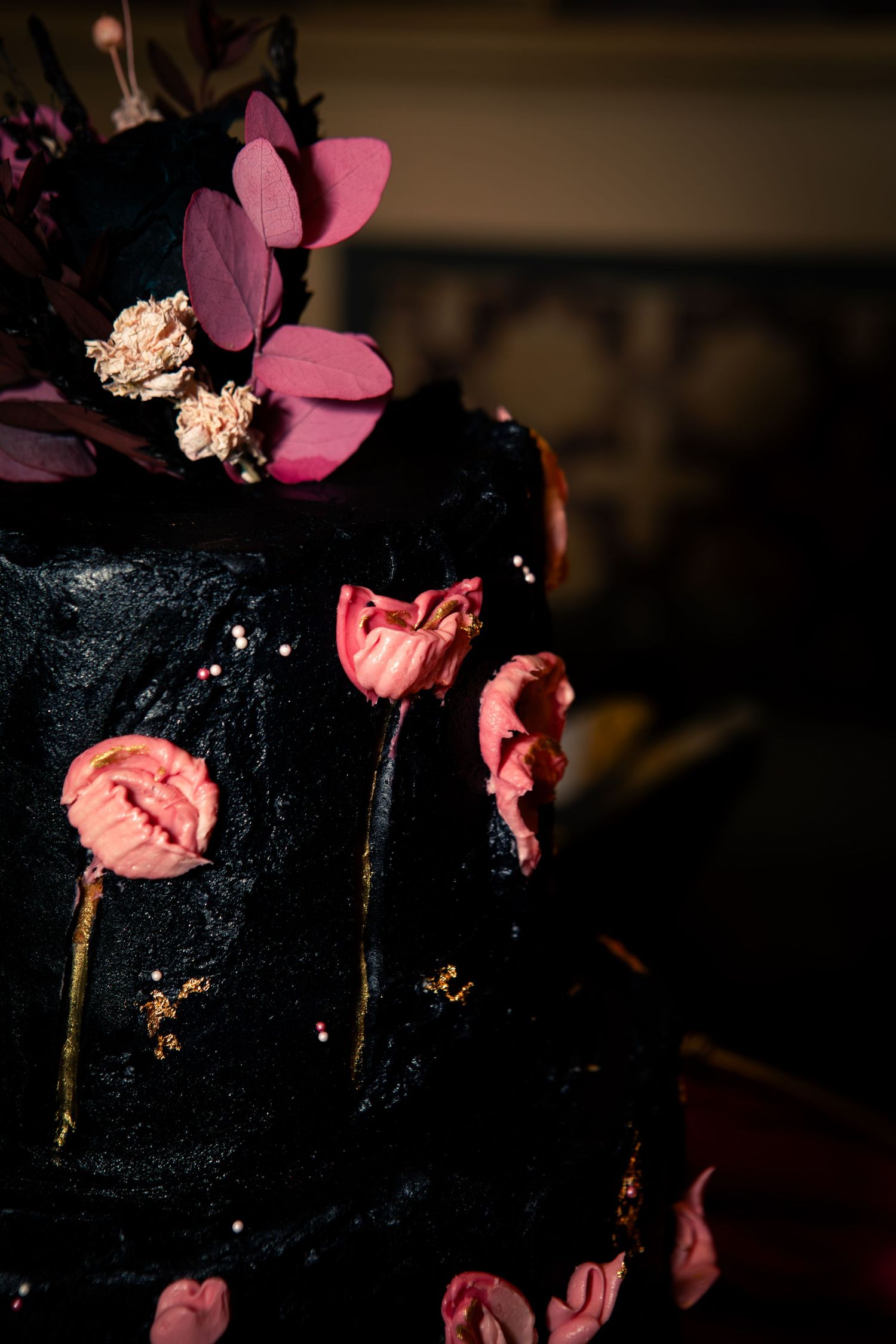 A dark black cake decorated with pink flowers and delicate petals against a moody background.