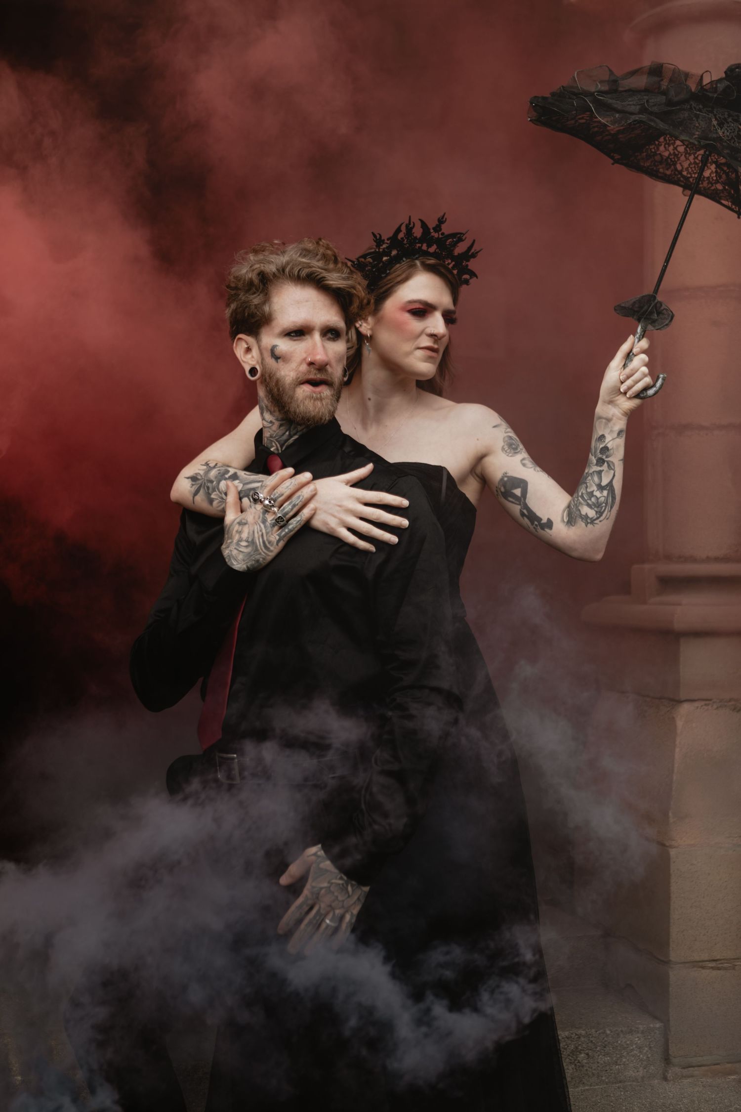 Two figures in dramatic poses surrounded by dark smoky atmosphere against red-tinted background for gothic photoshoot.