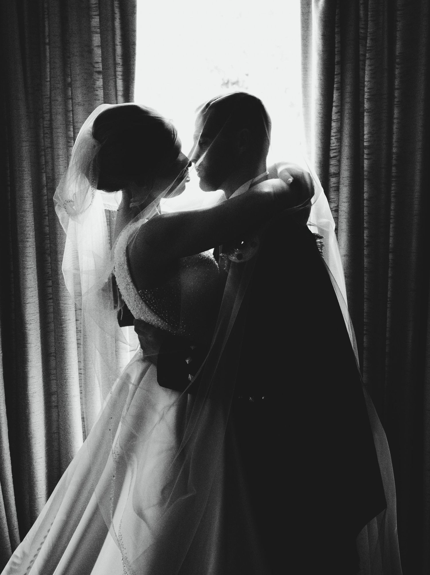 Silhouette of a romantic couple in wedding attire embracing by a window with dramatic backlighting.