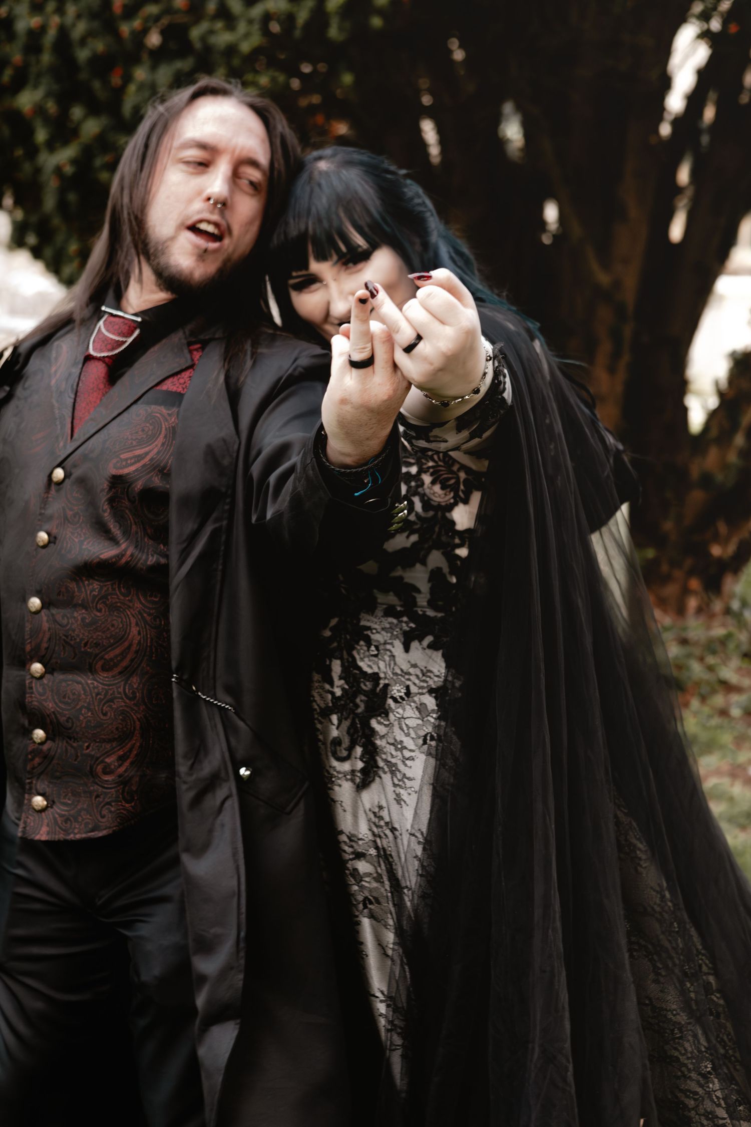 Gothic couple in Victorian-inspired attire showing ring in outdoor setting.