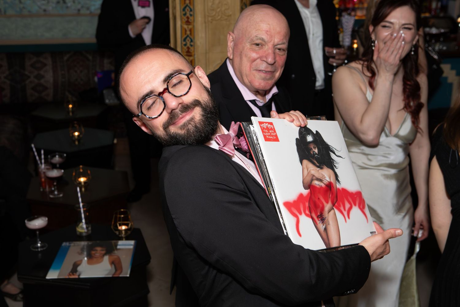 A man in glasses hugs a magazine at an indoor celebration event with guests in the background.