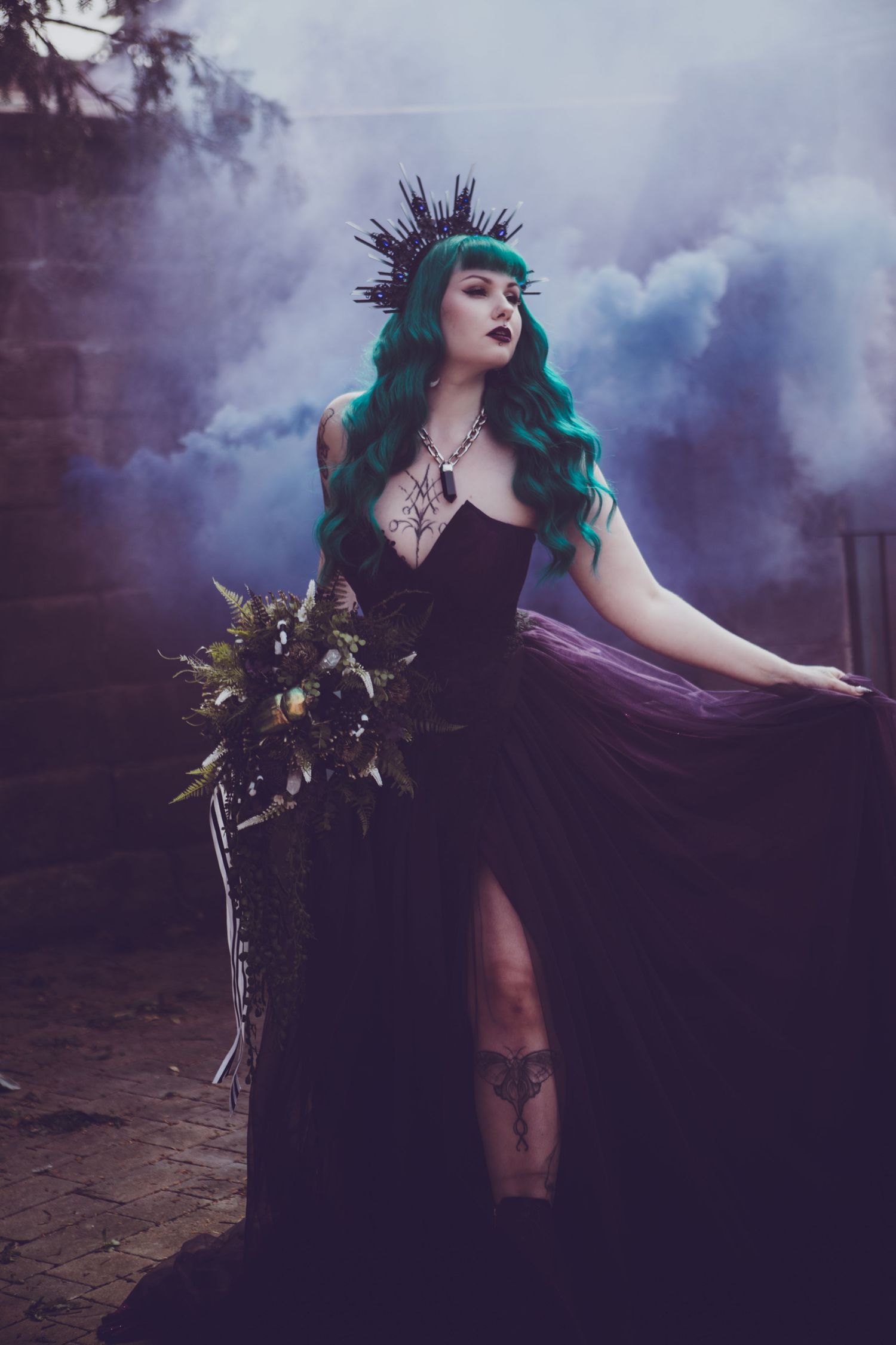 Gothic fantasy portrait with teal hair, spiked crown, dark dress and bouquet in moody purple smoke setting.
