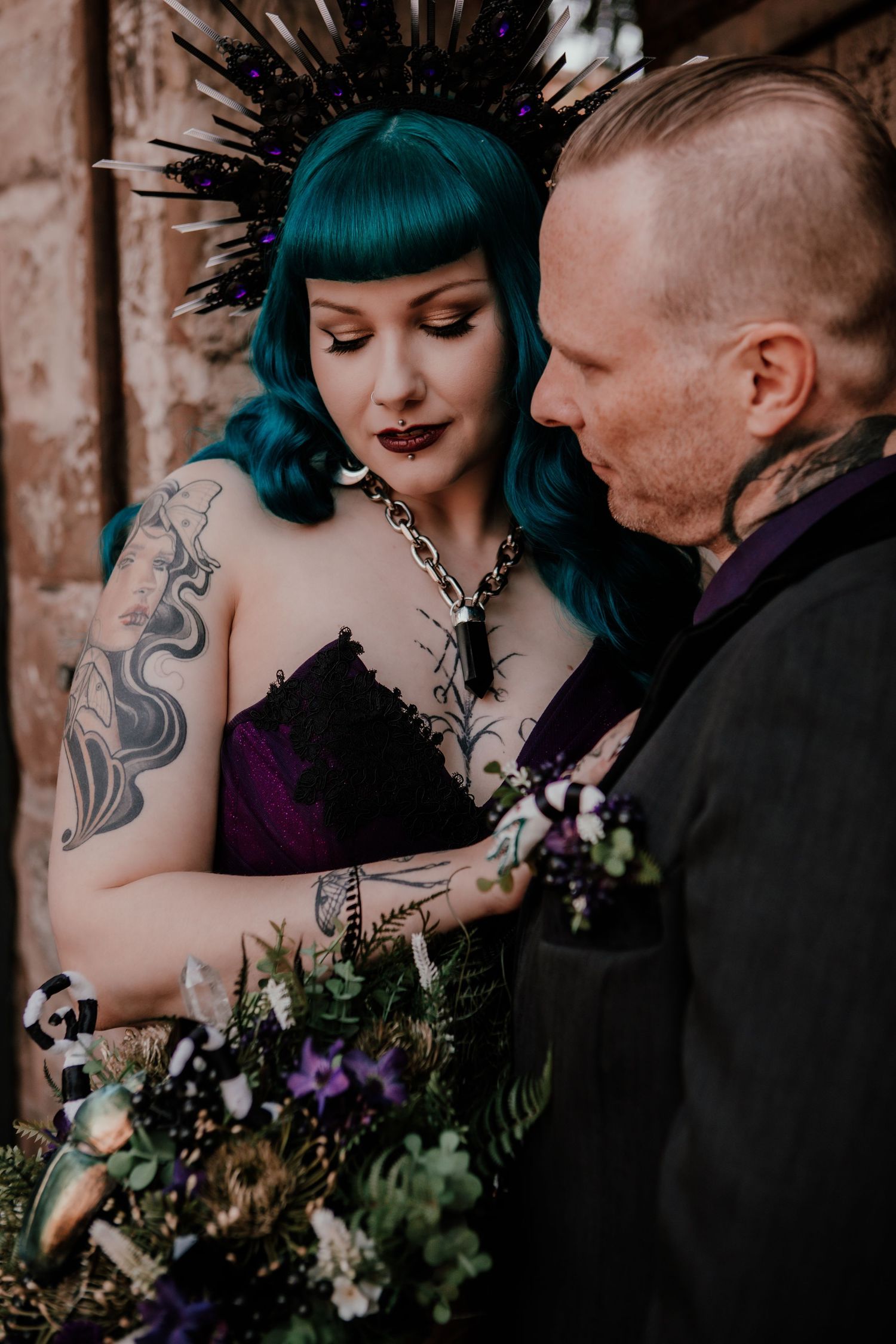 Alternative wedding couple in gothic style with teal hair, tattoos, and moody purple floral bouquet posing together.