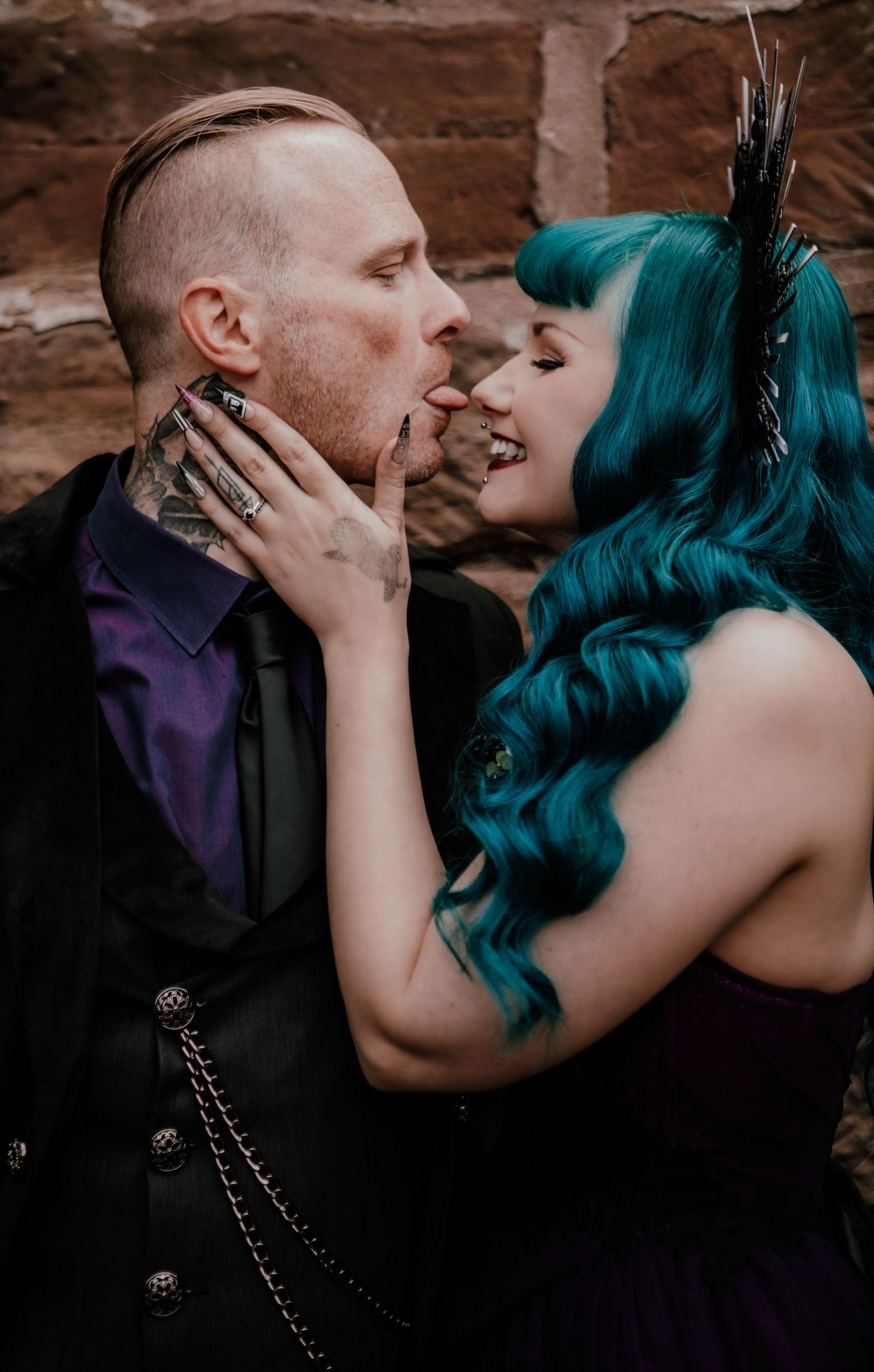 A Gothic-styled couple shares an intimate moment against a brick wall, with one wearing vibrant teal hair and black feathers.