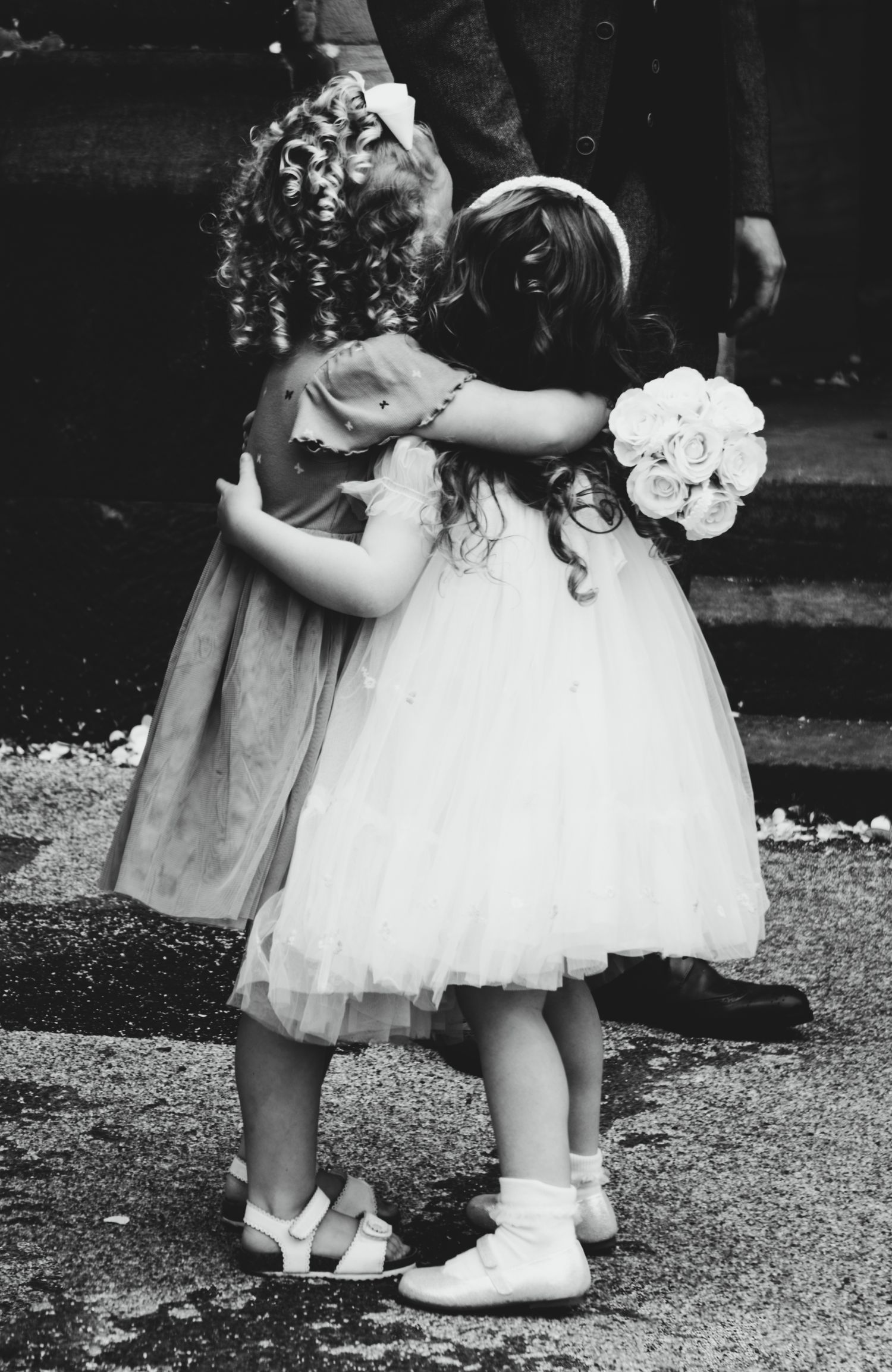 Two young flower girls in white dresses and curled hair share a sweet embrace at a wedding ceremony.