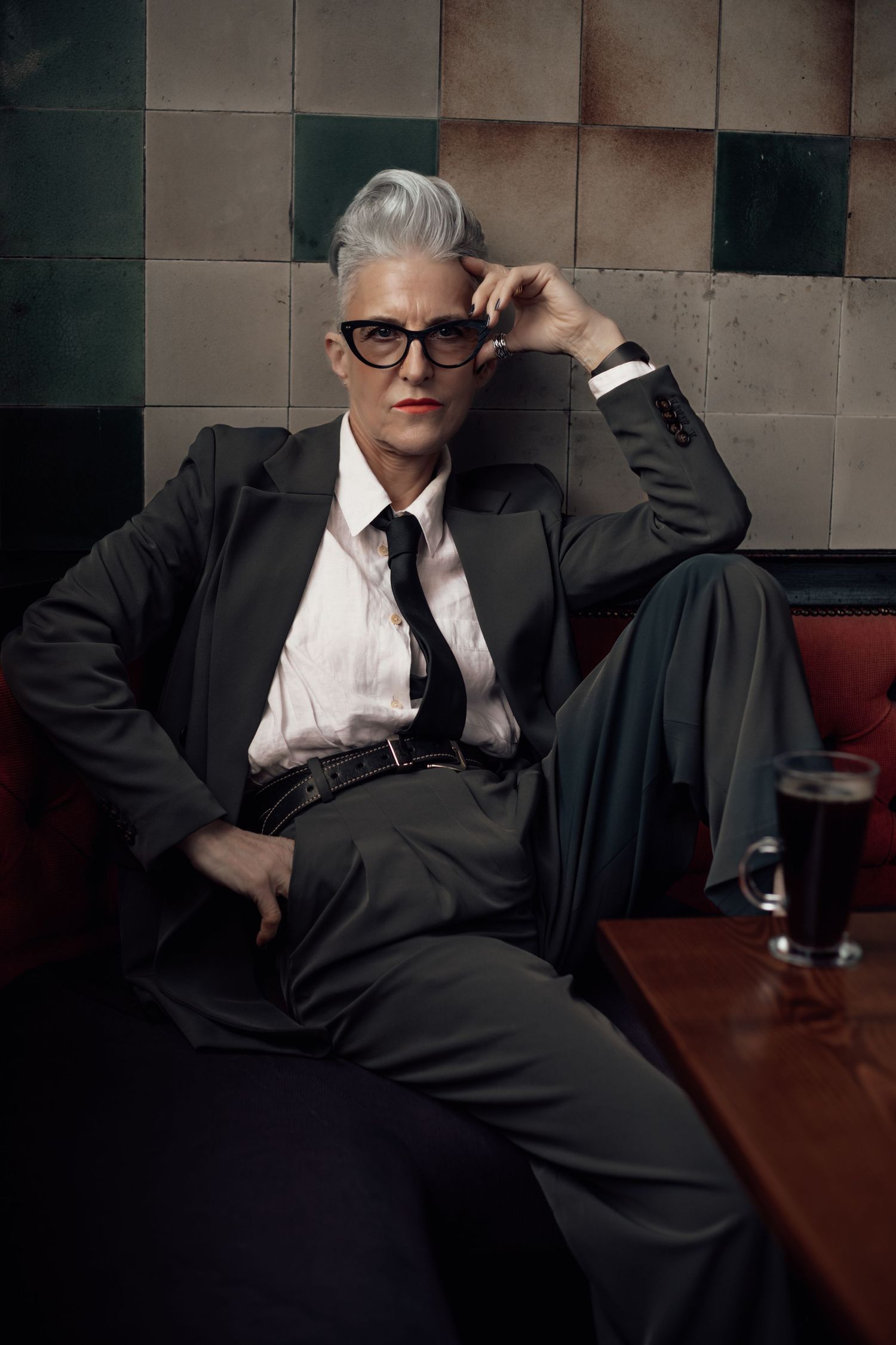 A person in business attire slouches at a table with a dark beverage in a dimly lit venue.