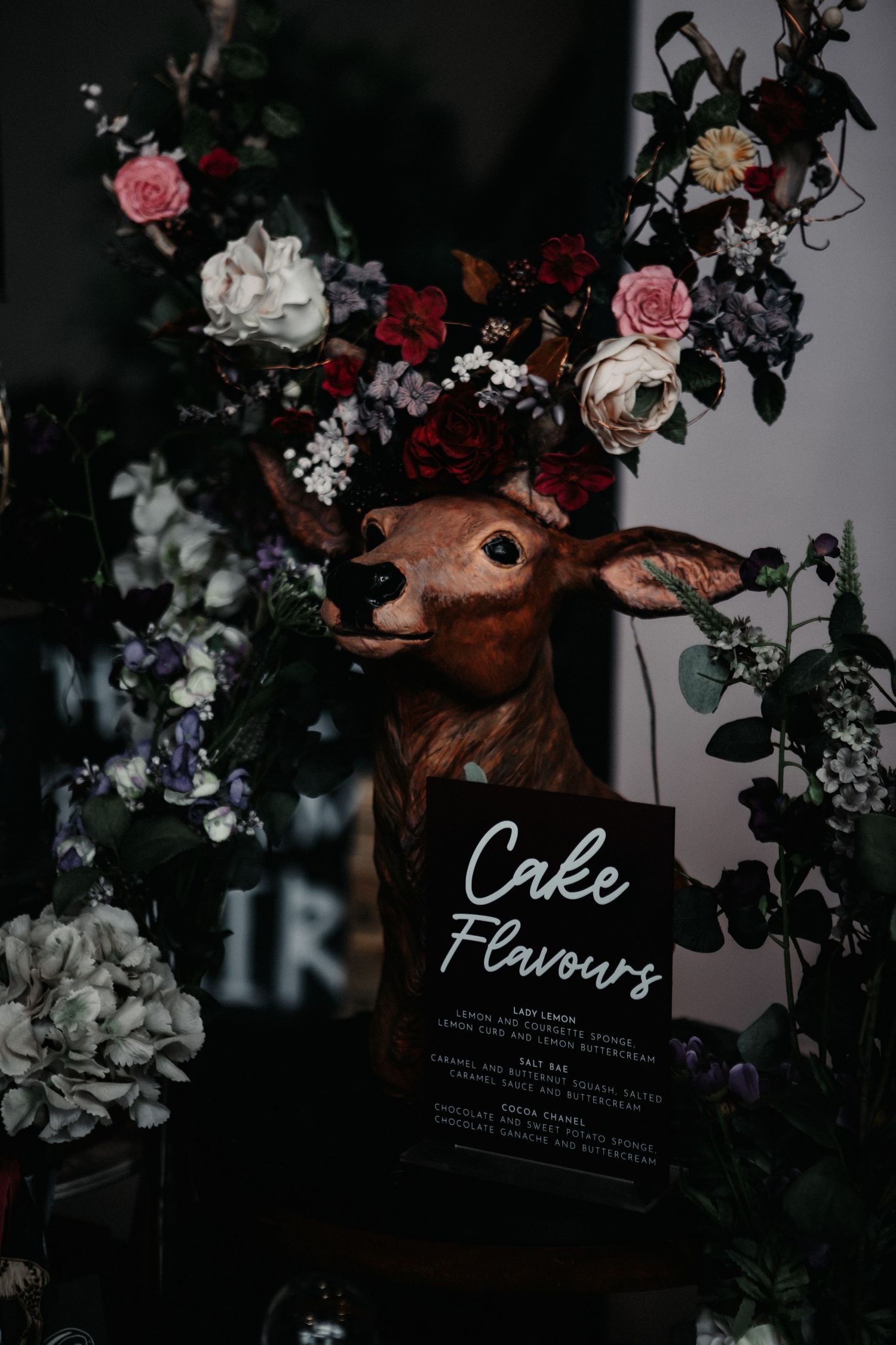 Dark moody floral arrangement with menu cards displaying 'Cake' and 'Flavours' text amid roses and greenery.