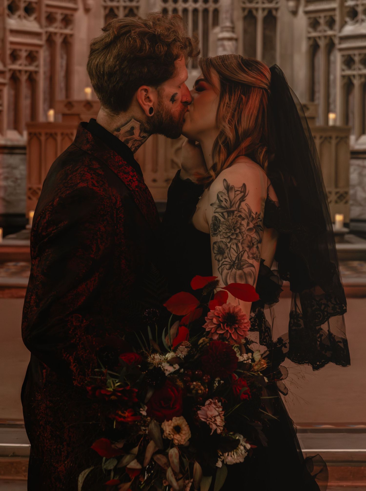 Gothic wedding couple kissing in front of ornate church architecture with red bouquet.