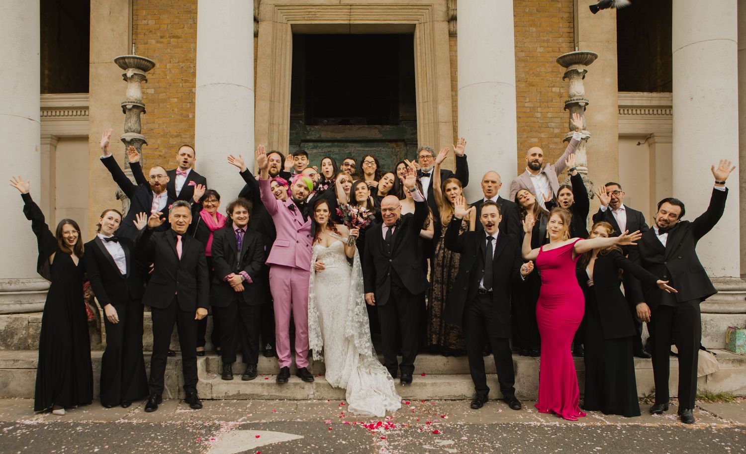 Large wedding party poses together in a formal lineup outside a stone building in formal attire for a portrait photo.