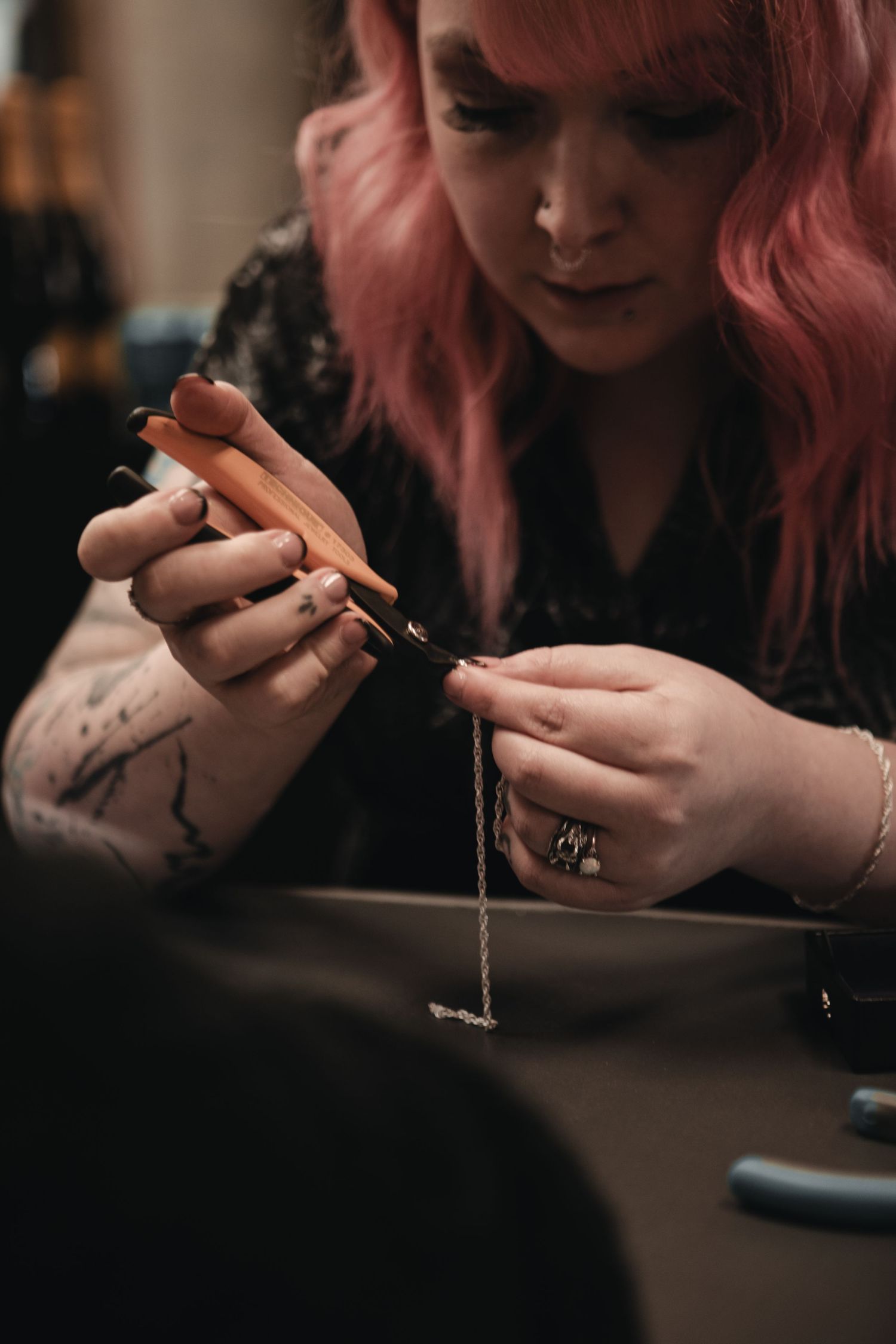 A person with pink hair works on jewelry making with focused attention while wearing black attire.