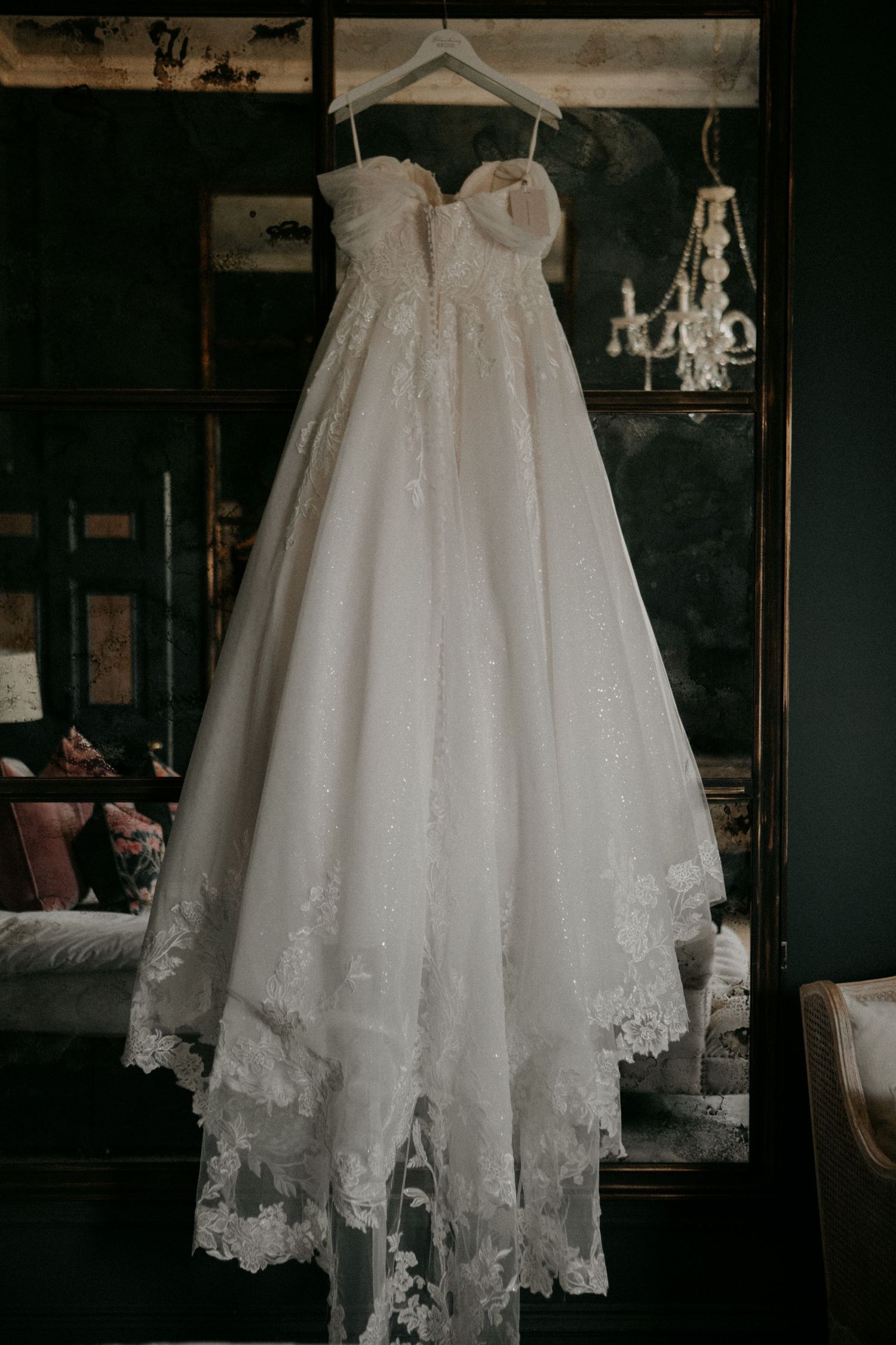 A elegant white wedding dress with lace details hangs on display against a dark wall with a chandelier in the background.