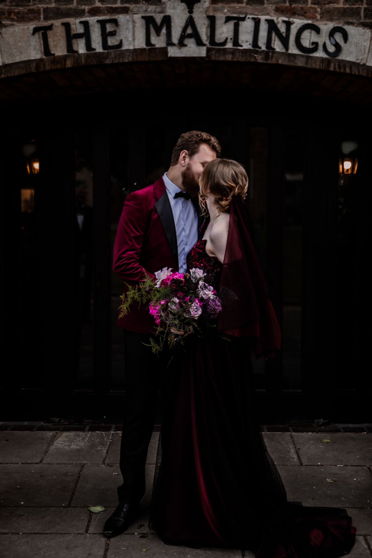 Romantic wedding photo under 'The Maltings' sign with burgundy velvet jacket and black dress holding vibrant floral bouquet.